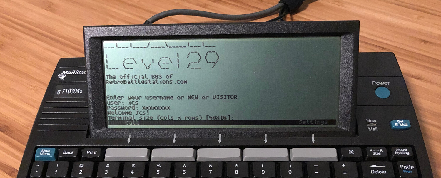 A black device with a monochrome LCD sits on a wooden table. It's keyboard extends below the frame. On the screen is the "Level 29" BBS service login.
