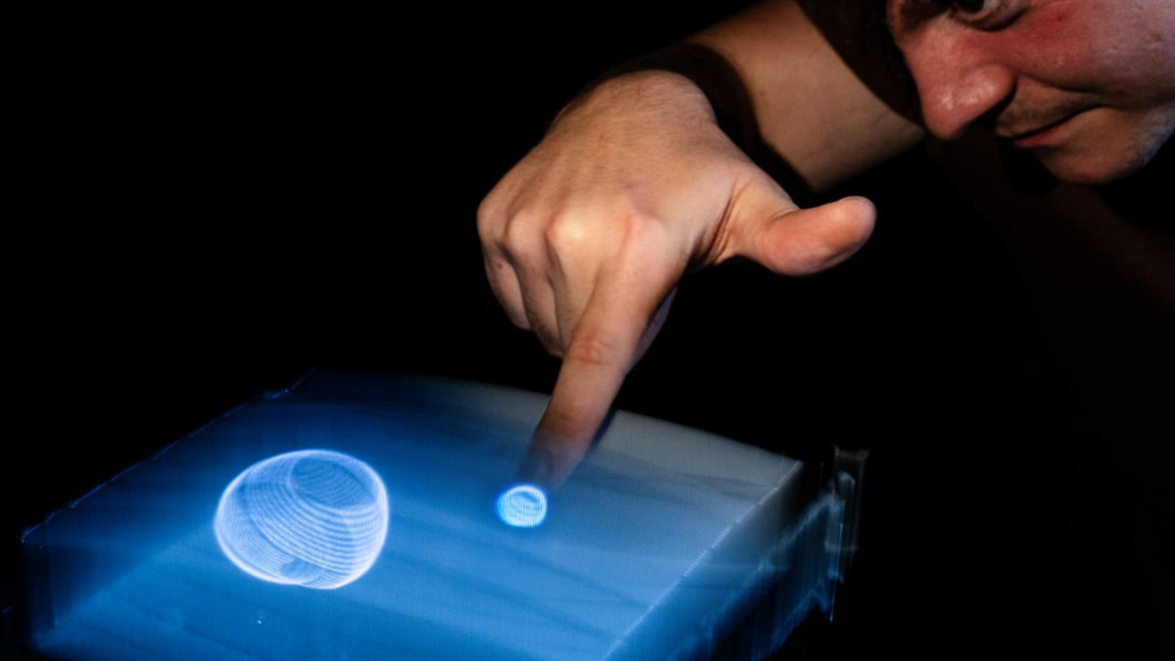 A man is looking at a volumetric display while using one finger to interact with it. Two roughly-spherical blue shapes are visible in the display, and he is moving his index finger toward one of them.