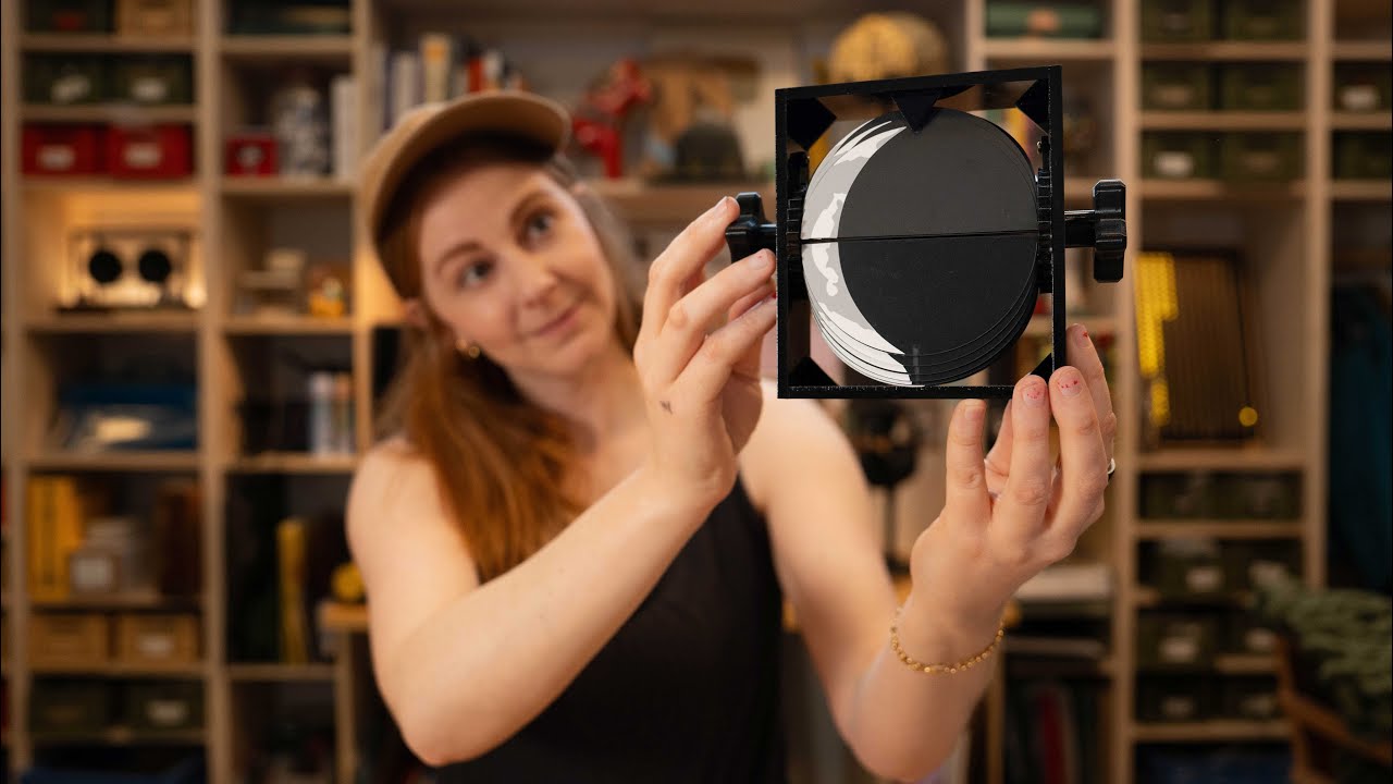 A woman in a ball cap and black tank top holds a black and white image of the Moon printed on black acrylic. It can fold and is mounted on a black plastic mechanism with turning knobs affixed to the ends. There are out-of-focus shelves in the background with various items and books on them.