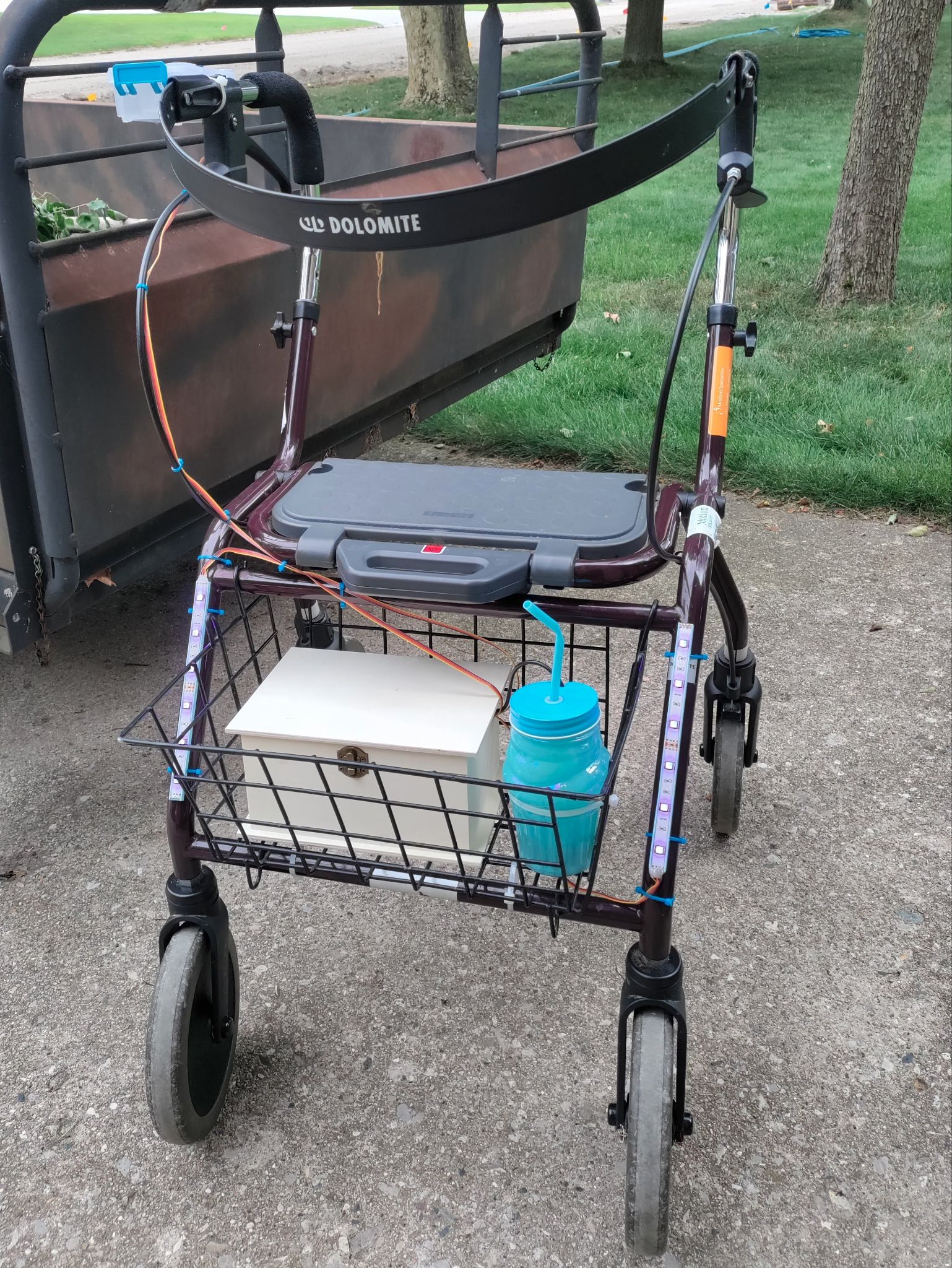 A walker with wheels and a plastic seat. In a basket under the seat is a white box with a brass latch. A blue water bottle sits next to it. Wires come out of the box and go up to the handles of the walker and to the water bottle (actually a water gun) and RGB lighting on the front of the walker.