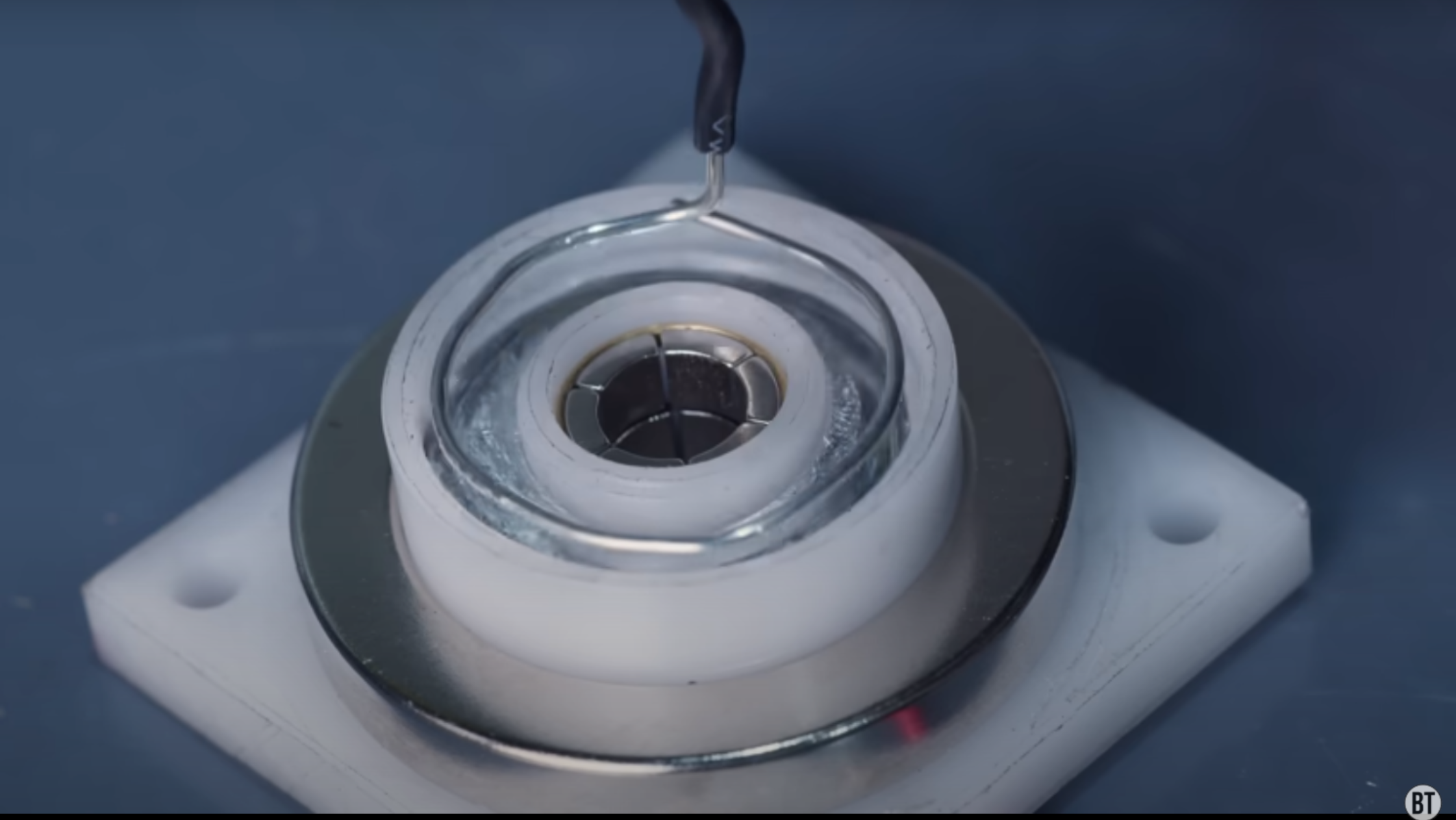 Two rings of magnets are shown encasing a circular channel in a white plastic piece. The channel is filled with liquid metal, and a loop of wire is about to be lowered into the metal.
