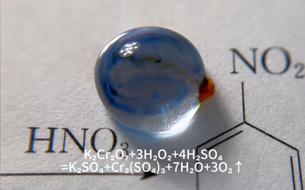 A picture of a single water droplet on top of what appears to be a page from a chemistry text. An orange particle is attached to the right side of the droplet and blue and black tendrils diffuse through the drop from it. Under the water drop, the caption tells us the reaction we're seeing is "K2Cr2O7+ 3H2O2 + 4H2SO4 = K2SO4+Cr2(SO4)3+7H2O+3O2(gas)"