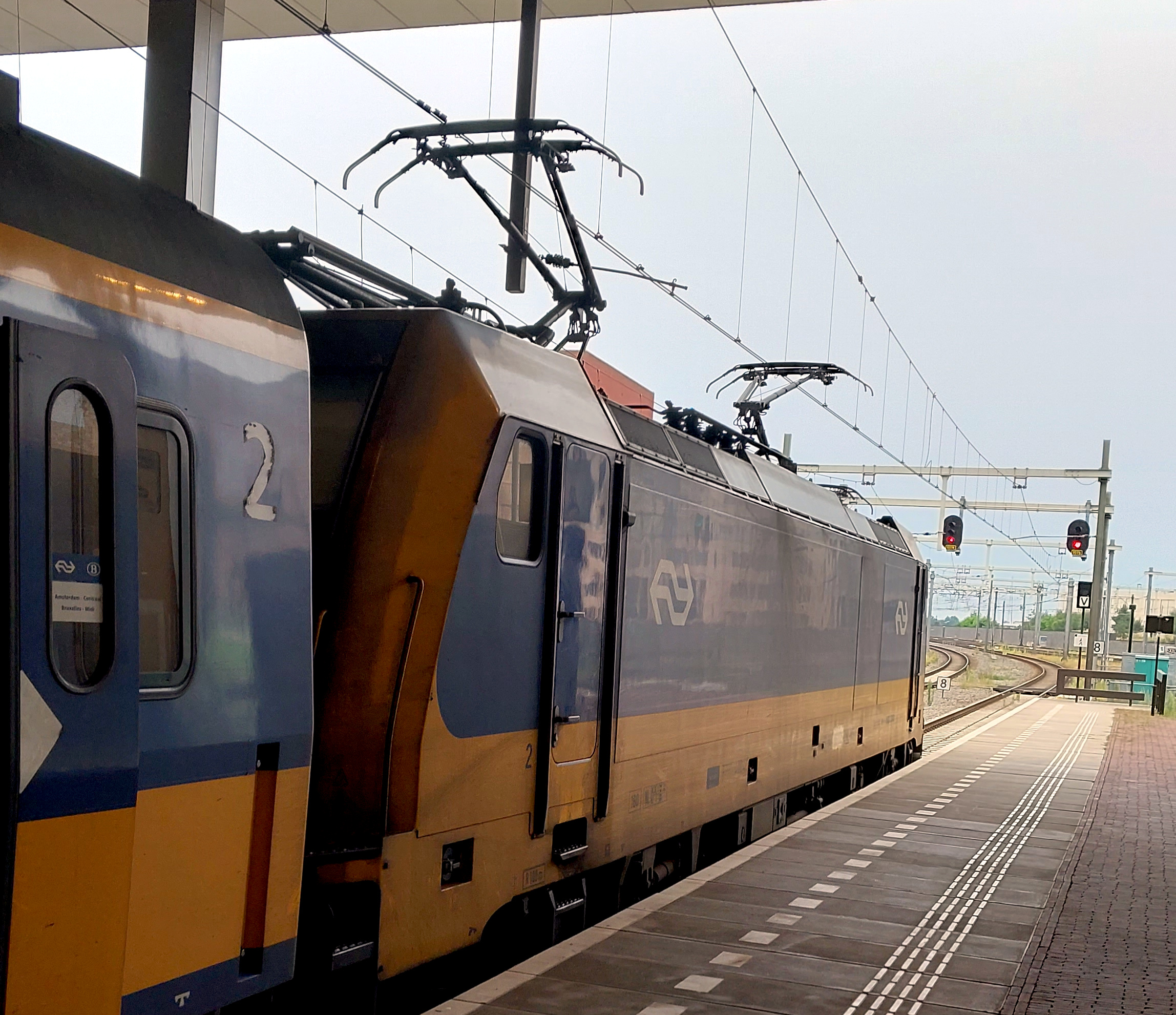 A blue and yellow electric locomotive at a station platform, pointing forwards towards some tracks which curve to the left in the distance.