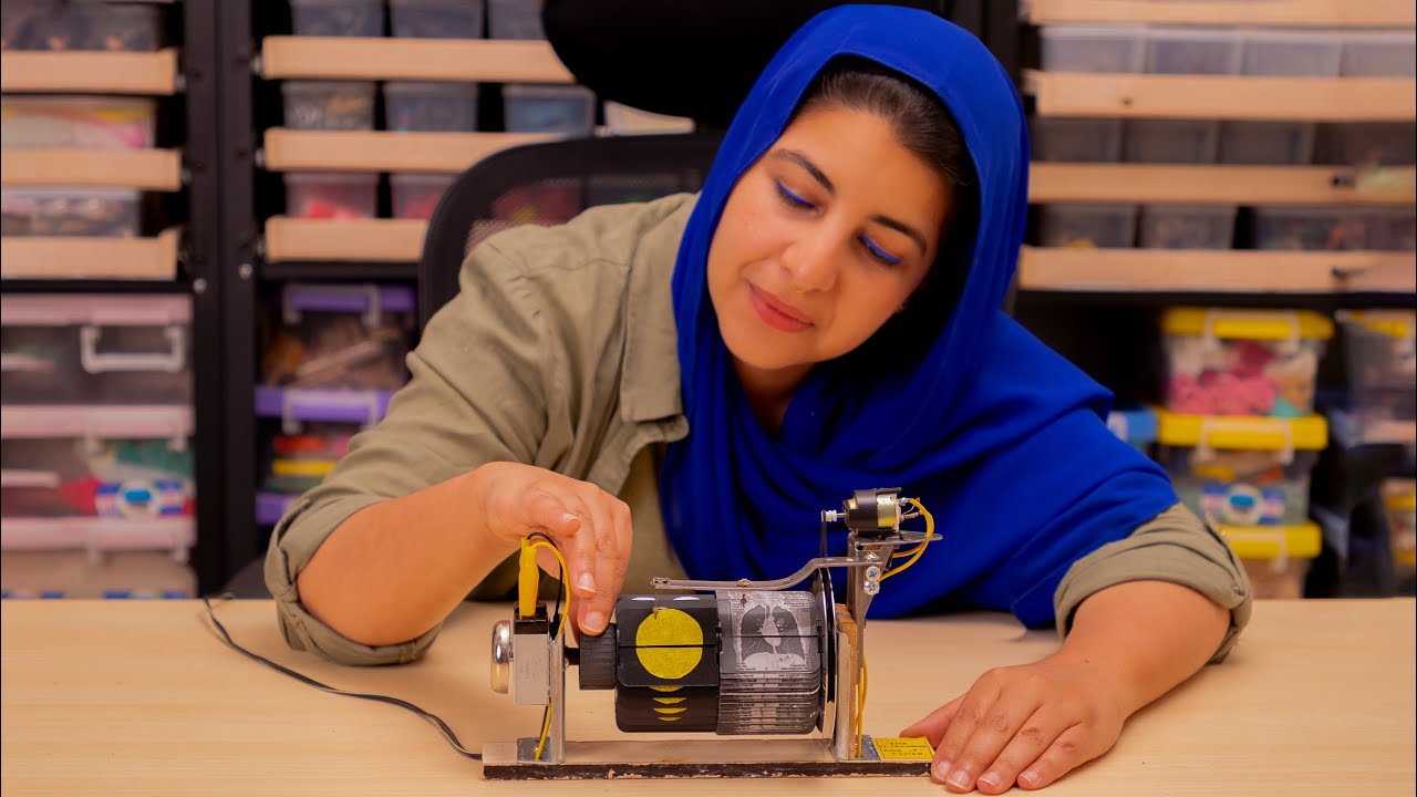 A woman in a richly-colored blue head scarf leans over a wooden table looking at the timer between her hands. The timer has a yellow circle on black flip panels on the left and black and white CT scans of a human torso on the right side. The frame is wood, and there is an electric motor on the upper right of the frame and a silver drum on the left of the frame beneath the woman's hand.