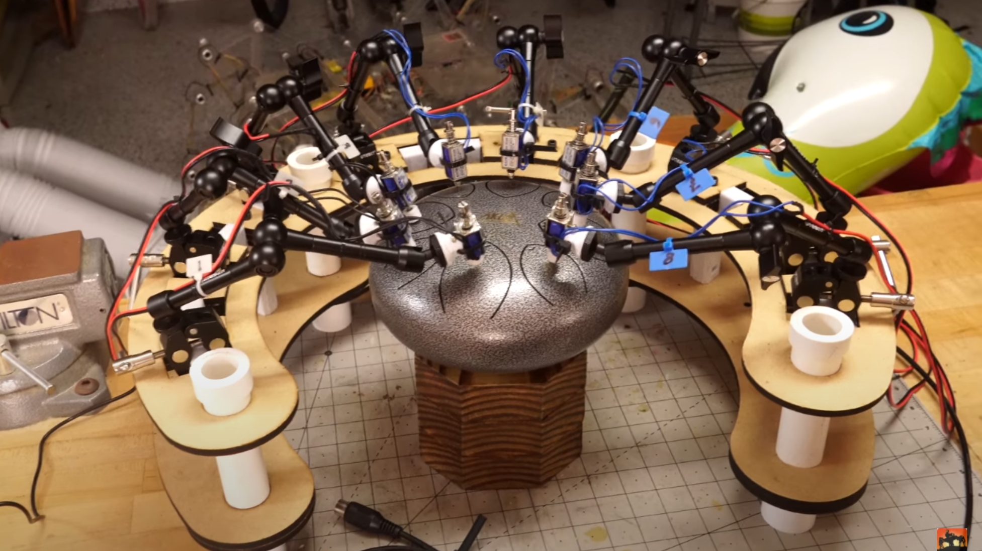 A C-shaped wooden frame is shown surrounding a circular tongue drum. The wooden frame holds eight black adjustable arms, at the ends of which are mounted solenoids, positioned just above the surface of the drum.