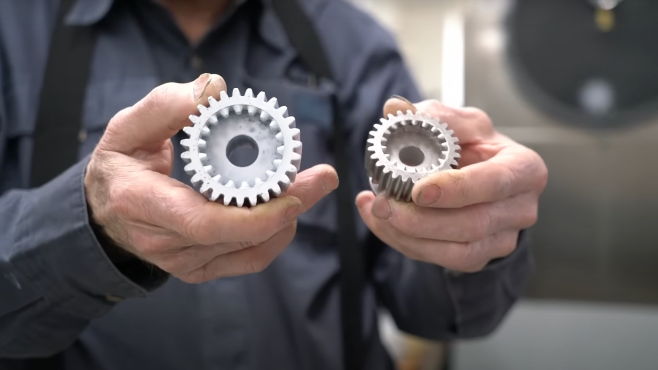 A man’s hands are shown in the foreground holding two splines, each with teeth both on the interior and exterior surfaces. Both are identical in shape, but one is larger and made of plastic, and the other is smaller and made of metal.