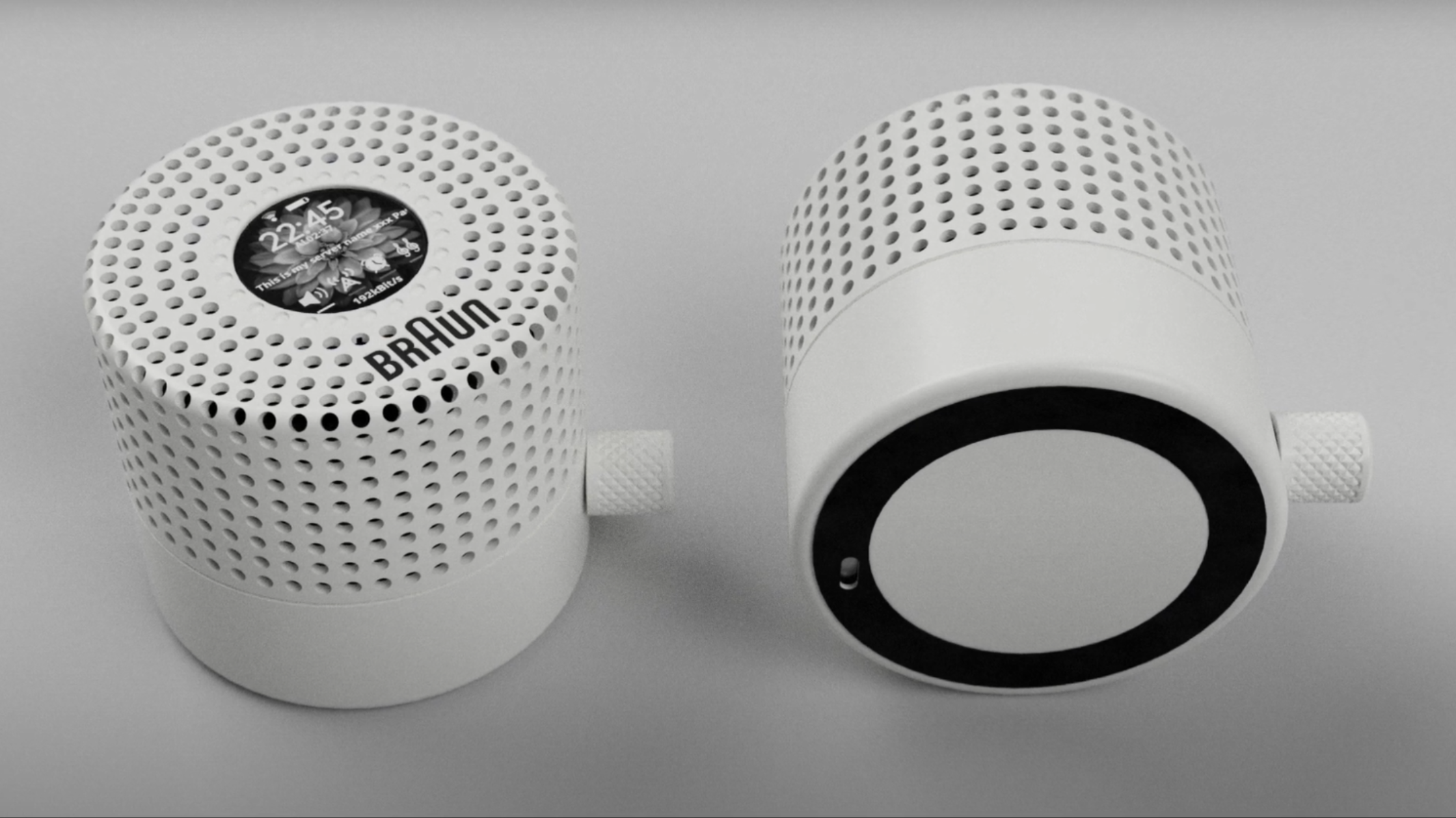 Two white, cylindrical speakers are shown. The sides and most of the tops of the speakers are covered in holes, and at the center of the top of each, there is a circular LCD display. The top of the speaker is marked “Braun.”