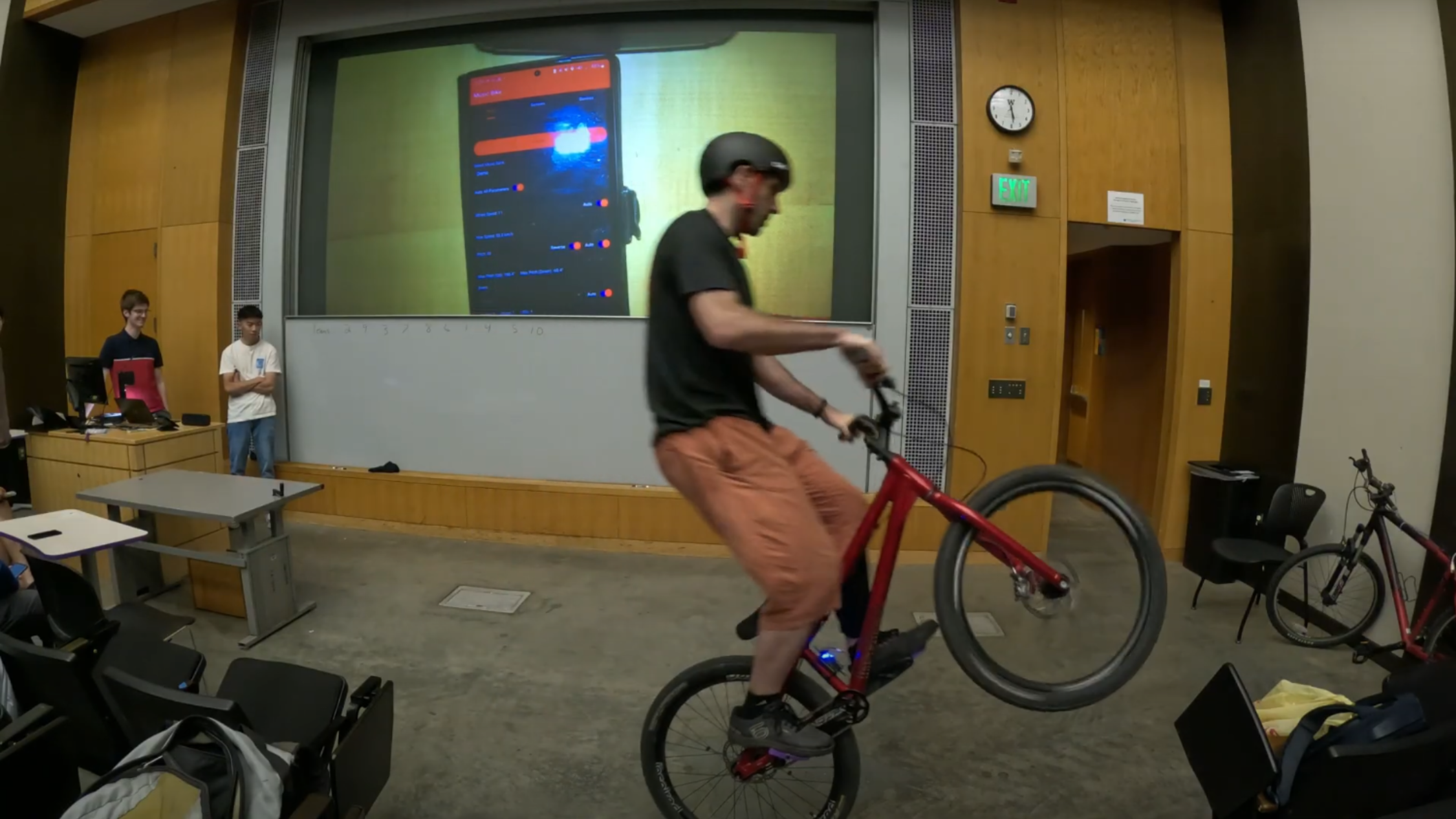 A man is shown performing a wheelie on a red bicycle in a classroom. In the background, a projector is displaying a phone screen running an indistinct app.