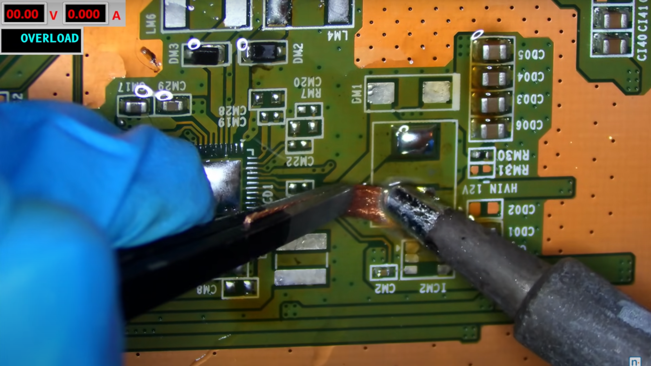 A circuit board is shown, with the tip of a soldering iron applied to a piece of solder wick positioned above a pad.
