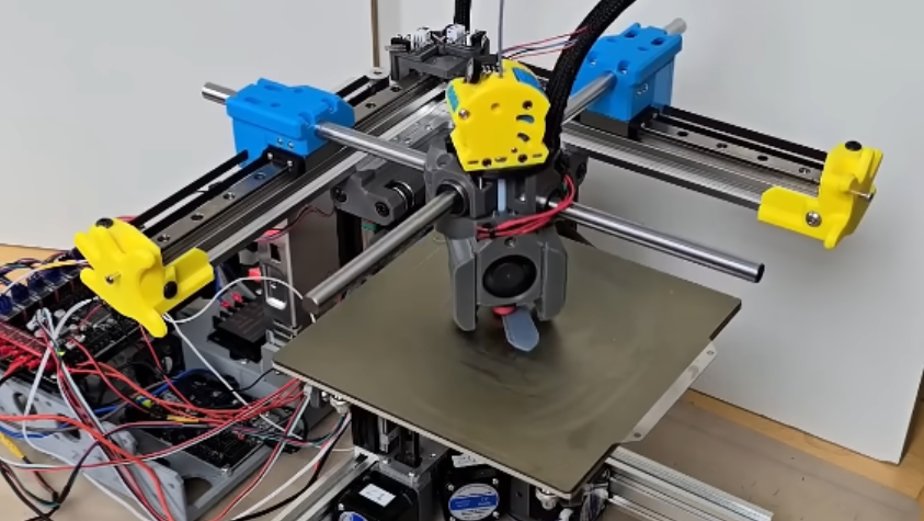 A 3D printer is in the process of printing a test piece. The printer has two horizontal linear rails at right angles to each other, with cylindrical metal rods mounted horizontally on the rails, so that the rods cross over the print bed. The print head slides along these rods.
