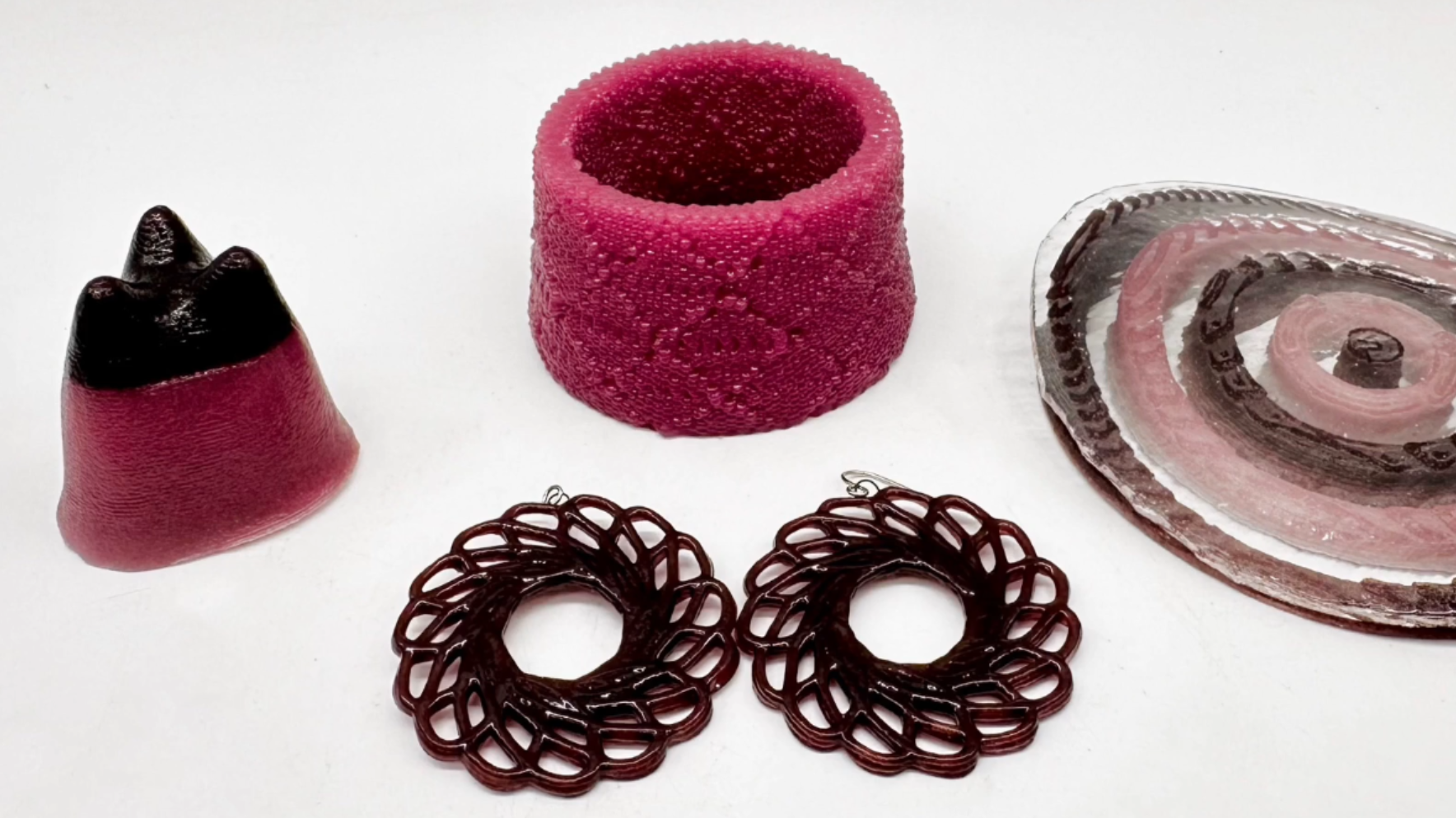 A variety of red and black glass objects are shown on a white background. In the foreground, there are two black spiral-patterned earrings. To the left is a red and black shape with three points on the top. On the right, a deformed glass sheet is shown bent over concentric red and black glass rings. In the center top is a red glass vase with a roughly-textured exterior.