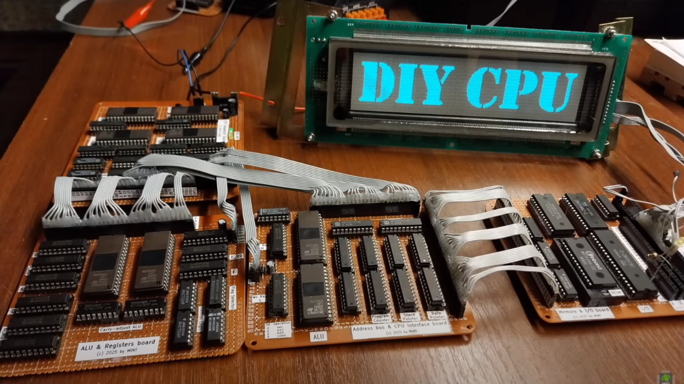 Four brown perf board circuits are visible in the foreground, each populated with many large DIP integrated circuits. The boards are connected with grey ribbon cable. Behind the boards a vacuum fluorescent display shows the words “DIY CPU.”