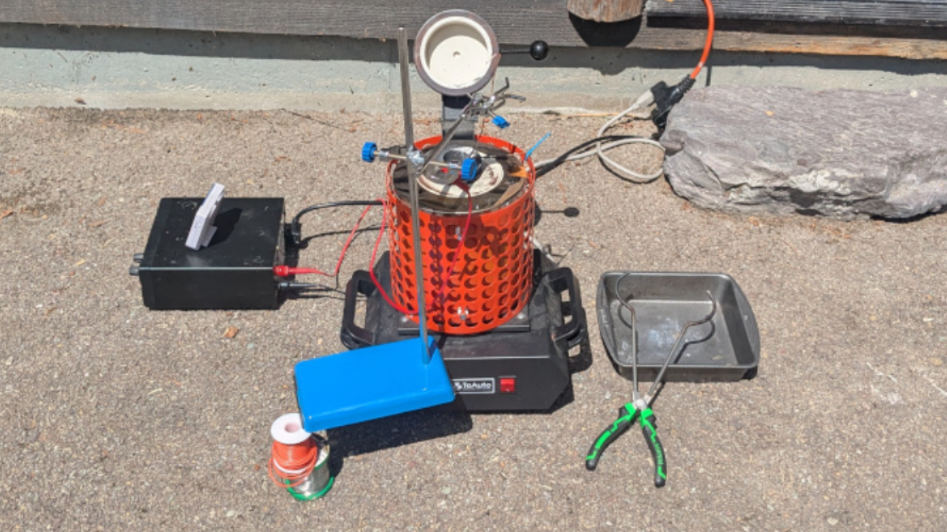 A cylindrical red furnace is in the center of the image. To the left of it is a black power supply. A stand is in front of the furnace, with an arm extending over the furnace. To the right of the furnace, a pair of green-handled crucible tongs sit on an aluminium pan.