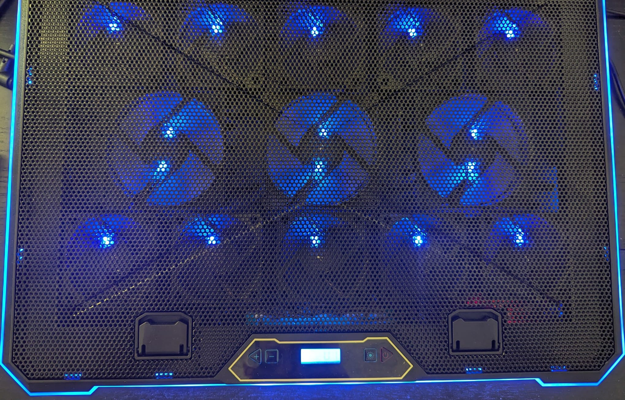 The top surface of a laptop cooler is visible. It consists of a black plastic mesh with thirteen fans visible behind it, with a blue backlit screen at the bottom of the cooler. There is blue LED backlighting behind each fan, and around the border of the cooler.