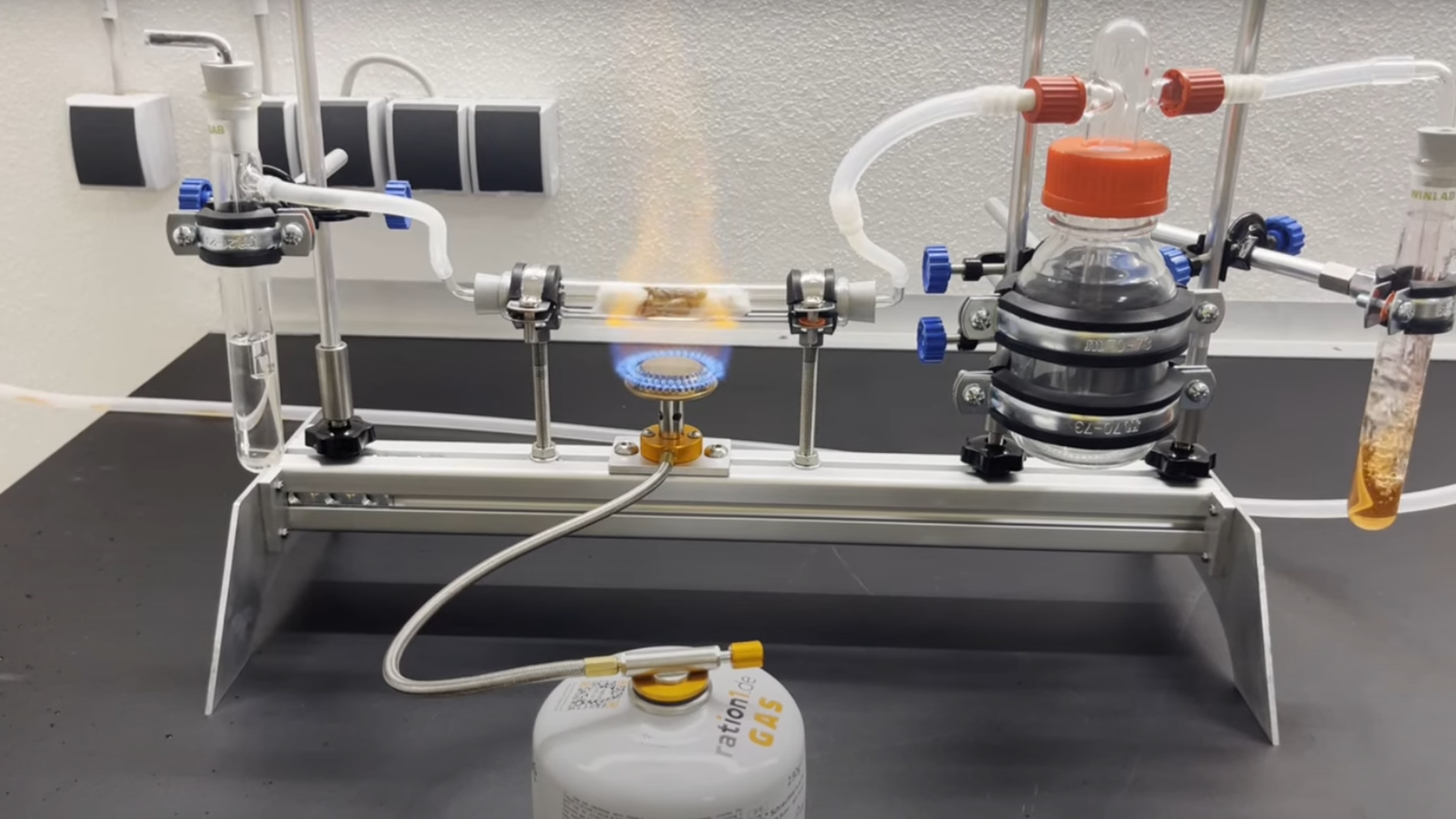 An aluminium frame is visible, supporting several connected pieces of chemistry equipment. At the left, there is a tube containing a clear solution, with a tube leading to a clear tube heated by a gas flame, with another tube leading to a clear bottle, which has a tube leading to a bubbling orange solution.