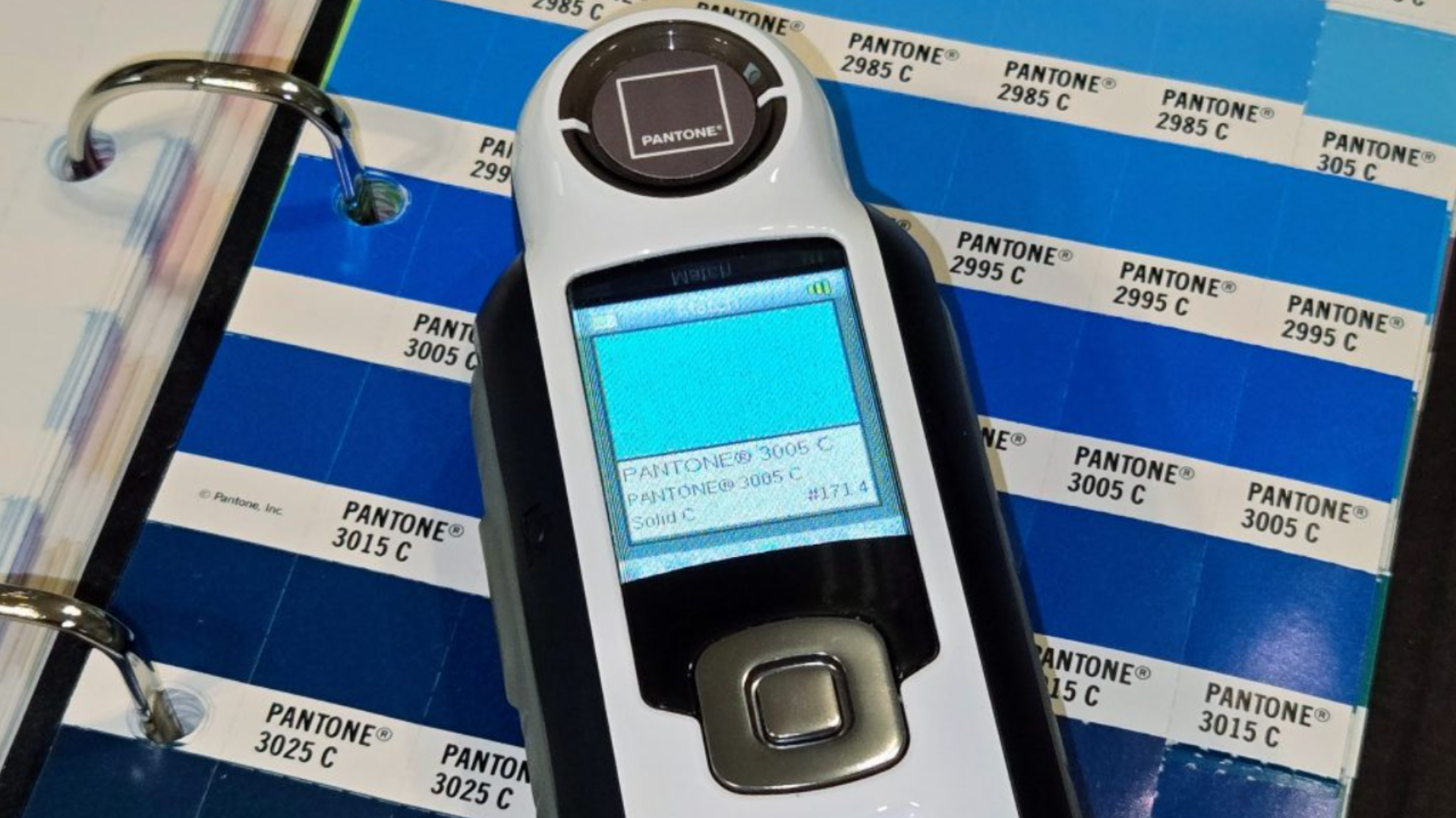 A long, rectangular electronic device is shown in front of a book of colour swatches. A small LCD display on the electronic device says “PANTONE 3005 C,” with additional color information given in smaller font below this.