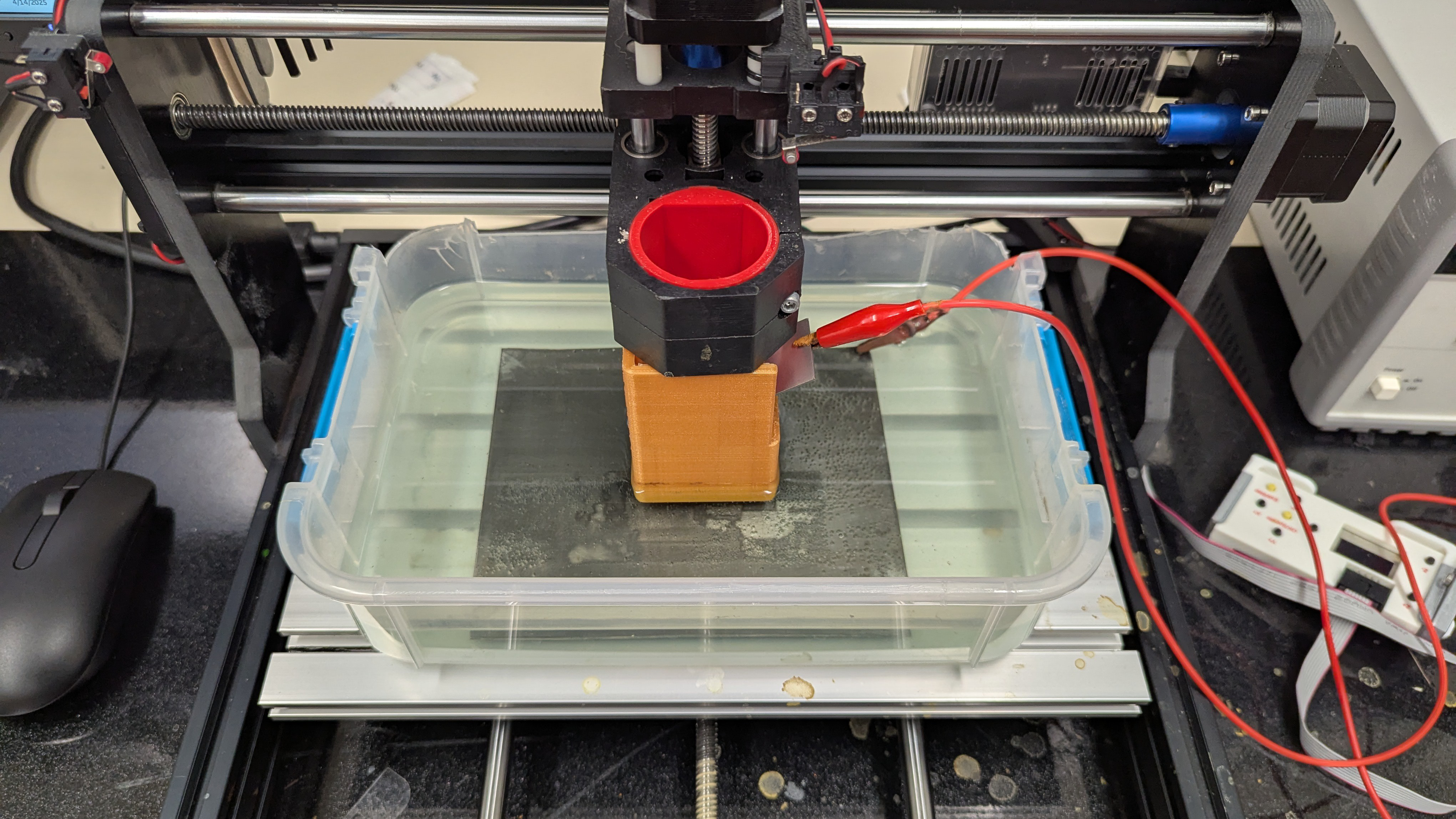 The bed of a small CNC machine is shown. A plastic tub is on the bed, and in the tub is a sheet of metal under a pale green solution. In place of the spindle of the CNC, there is a rectangular orange tube extending down into the solution. A red wire runs to this tube, and a black wire runs to the sheet of metal in the tub.