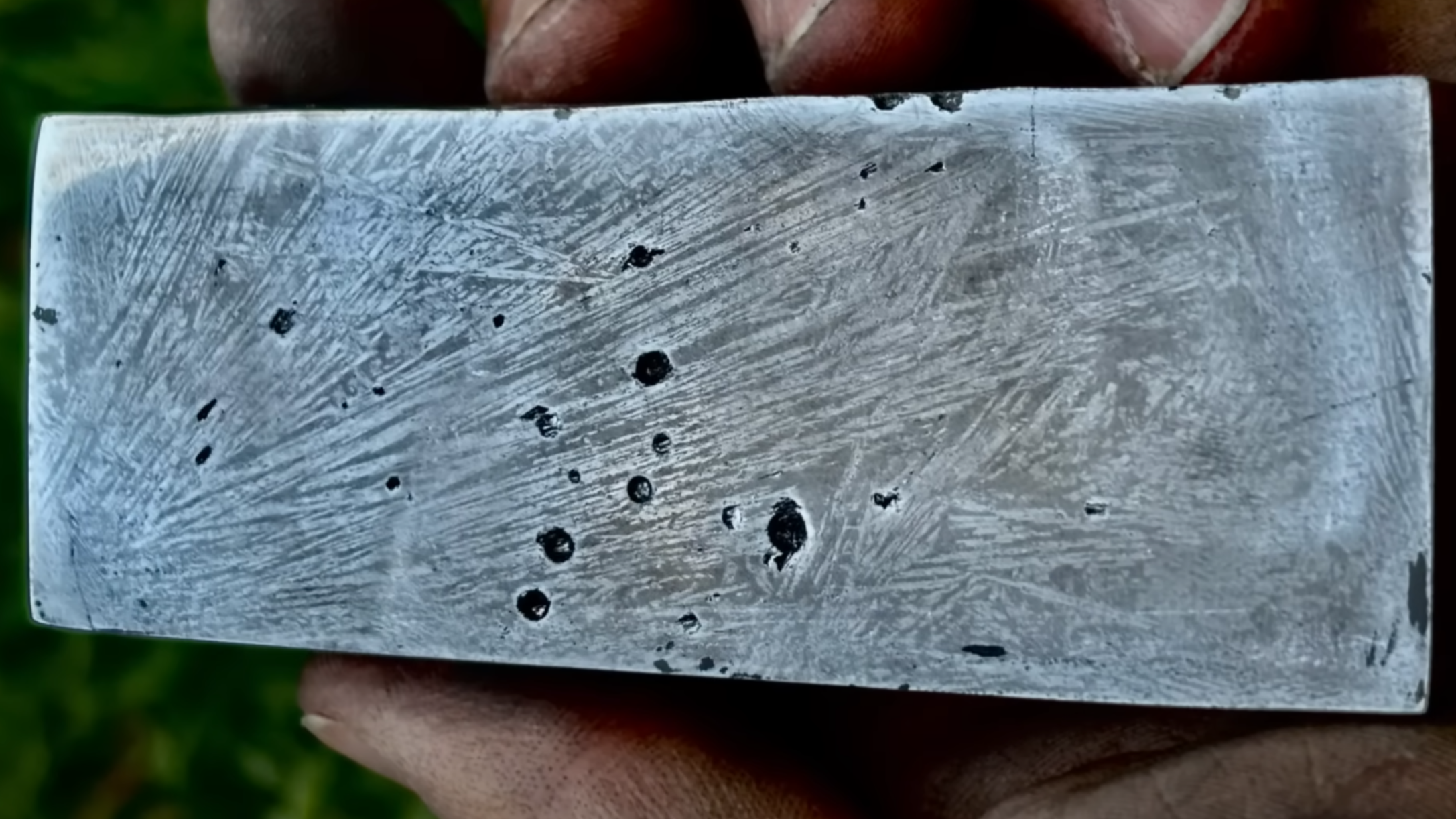 A man’s hand is shown holding a polished metal billet. The billet has a few voids in the surface, and the surface shows a pattern of lighter lines against the darker metal background.
