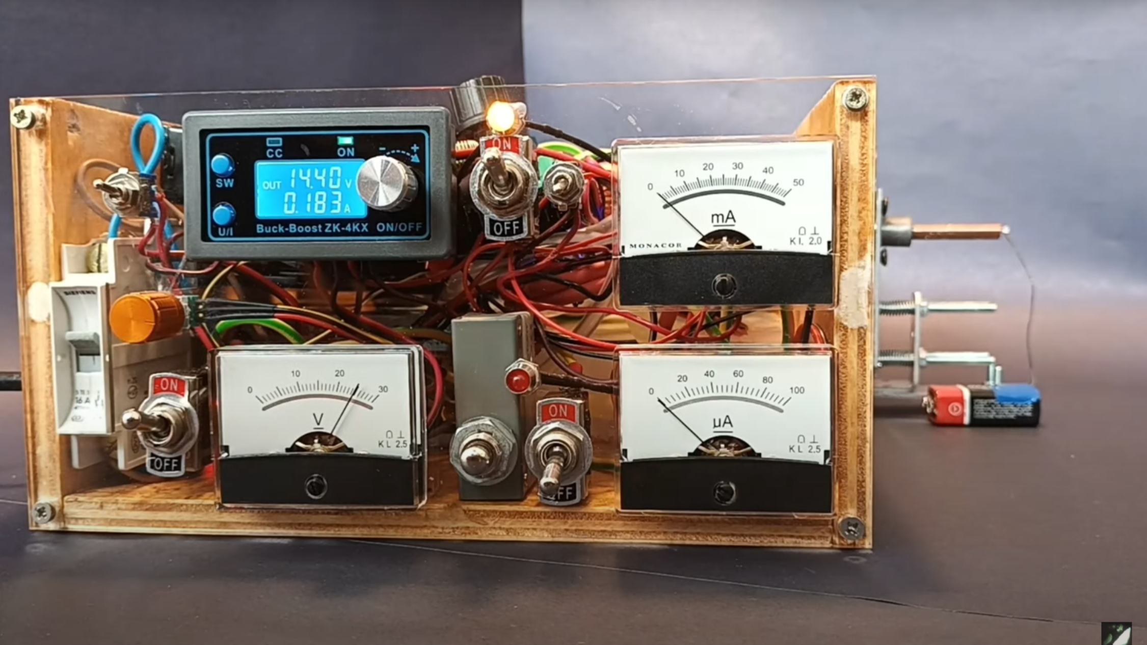 A plywood box with a clear plastic front is shown. Three needle gauges are visible on the front of the box, as well as a digital display, several switches, and some indicator lights. At the right of the box, a short copper tube extends from the box.