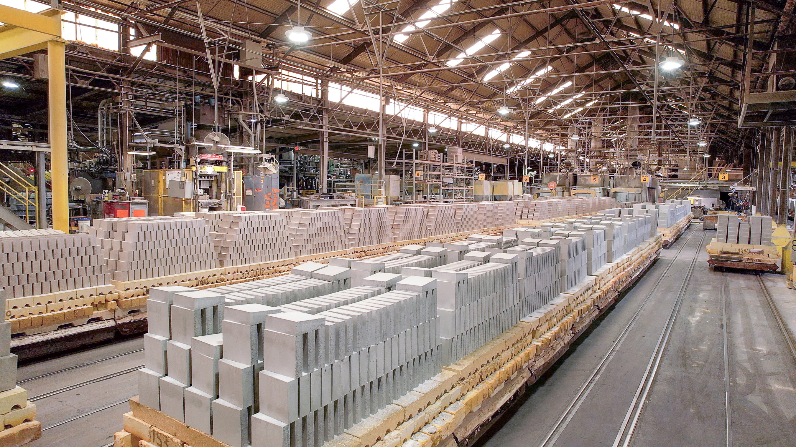 A photo of a large warehouse with many skylights and windows near the roof. In the middle of the image extending out into the distance are hundreds of grey refractory bricks stacked on top of a smaller set of brown bricks stacked on top of pallets. There appear to be rails on the floor of the warehouse and small dollies underneath the pallets.