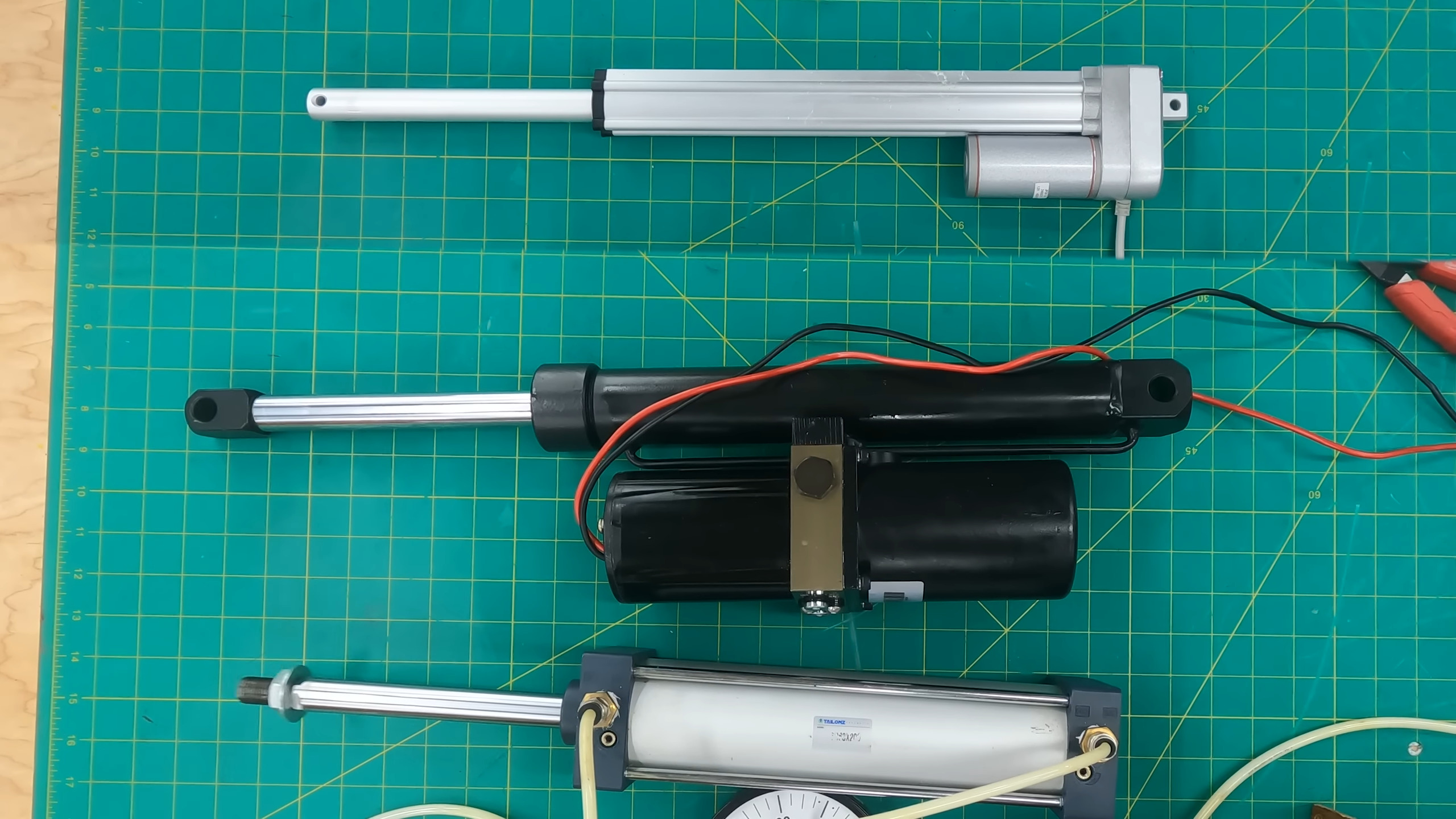 A set of three linear actuators set atop a green with yellow grid cutting mat. The electric actuator on the top of the image is silver and has a squarish tube. It is slender compared to the other two. A black, hydraulic actuator sits in the middle and is the largest of the three. A silver pneumatic actuator at the bottom of the image is the middle sized unit.