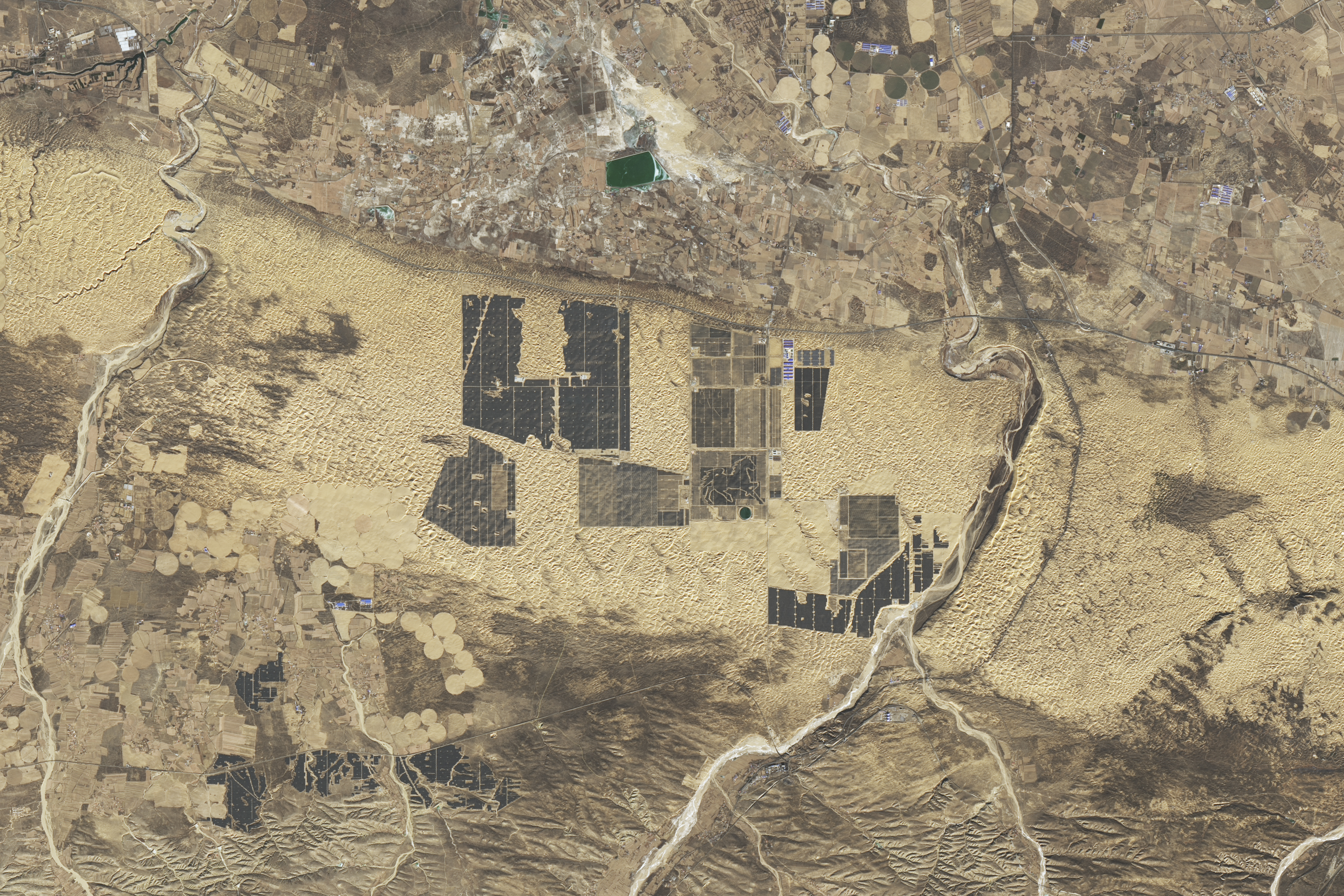 An overhead image of the Kubuqi Desert Great Solar Wall. It shows a series of clusters of bluish solar panels arranged throughout a light brown and dark brown desert. One of the arrays contains an image of a horse made of solar panels.
