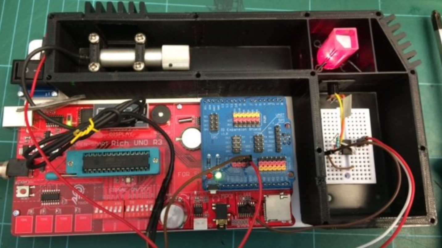 A 3D-printed case encloses a number of electronic components. In the top left of the case, a laser diode is mounted. In the top right, the laser beam is shining into a cuvette, which is glowing red from scattered light. In the bottom right, a small breadboard has an integrated circuit and a few parts mounted. In the bottom left is a large red circuit board marked “Rich UNO R3.”