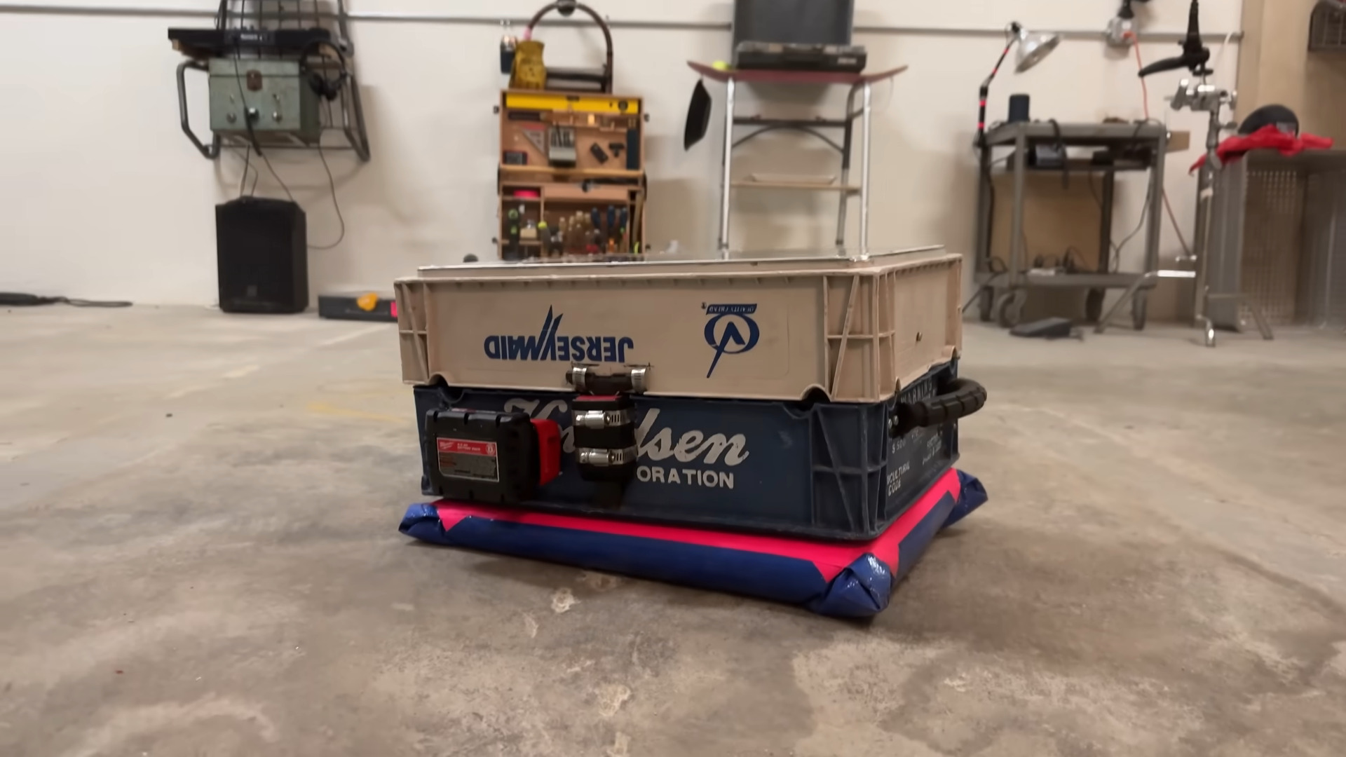 A suitcase made of two rectangular plastic crates latched together sits prone on a concrete floor. The top shell is a beige-ish grey with a navy check and the word "JerseyMaid" on it updside down. The navy bottom crate is navy with the letters "lsen" in large cursive font ant the letters "ORATION" in smaller print below it. Much of the text is covered by a large latch and a power tool battery mount. Bright pink tape affixes a blue tarp skirt to the bottom of the hover suitcase.