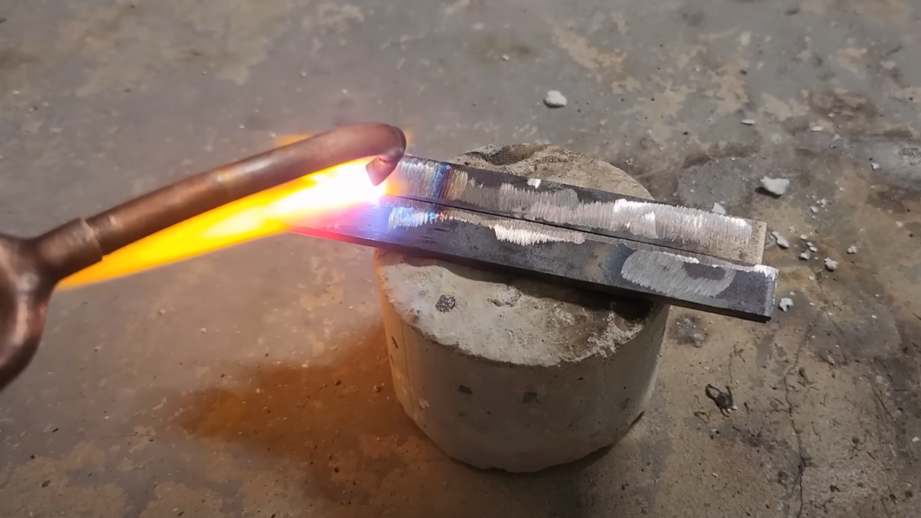 Two pieces of metal are sitting next to each other on a brick, with one edge touching. The copper end of a torch is directing a flame against the metal, and the metal is glowing brightly around the point where the flame contacts it.