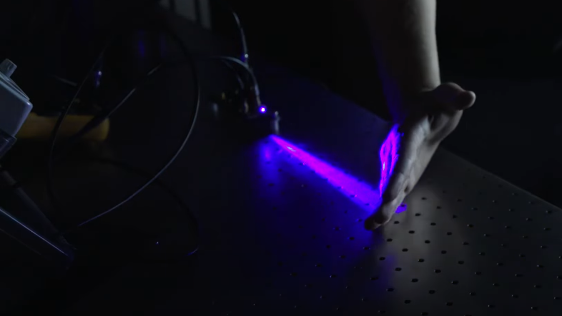 A violet laser beam is shown expanding outward from a diode in a darkened room and illuminating the back of a man's hand.