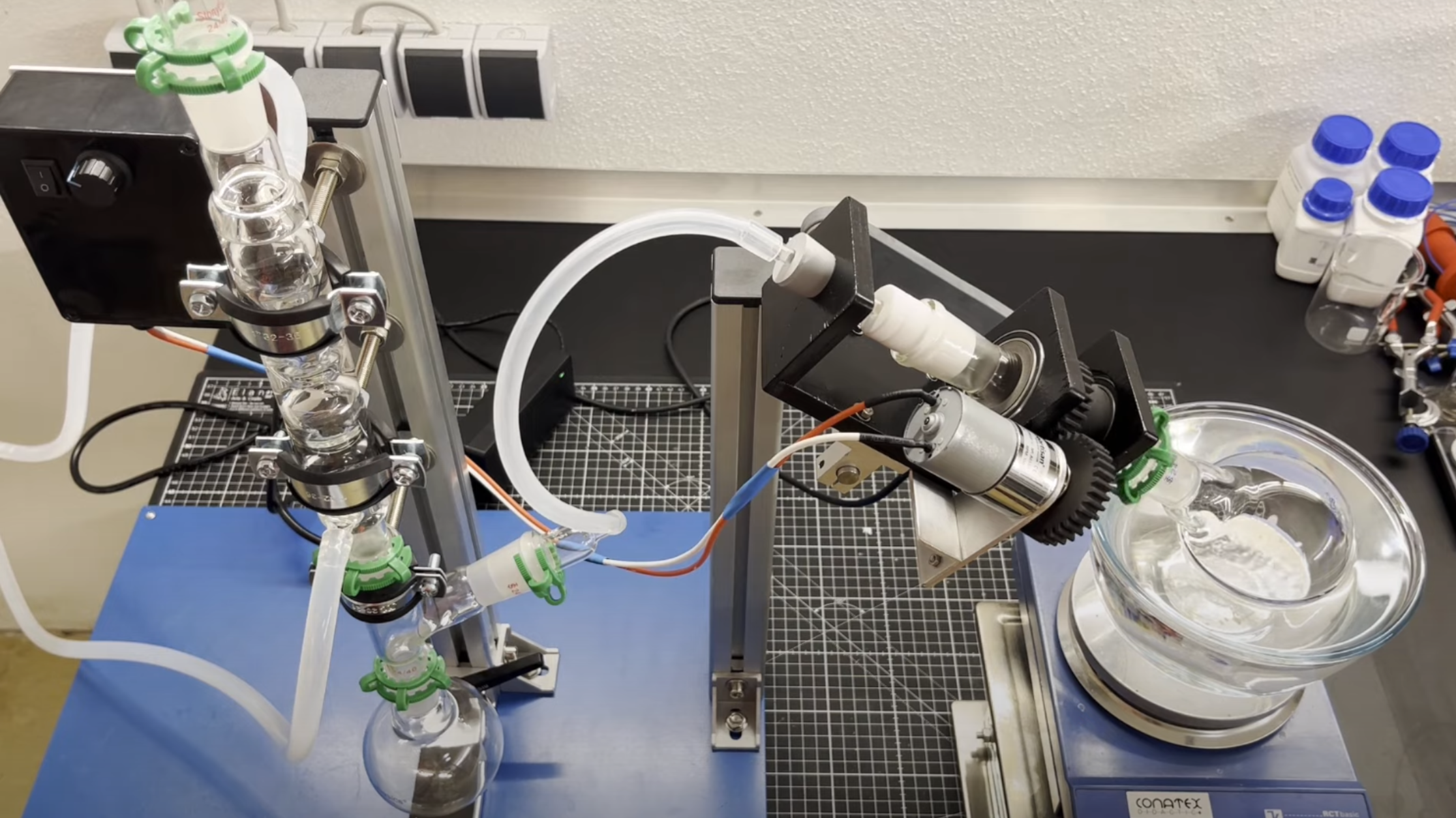 A laboratory benchtop is shown. To the left, there is a distillation column above a collecting flask, with a tube leading from the flask to an adapter. The adapter has a frame holding a glass tube with a teflon stopper at one end, into which a smaller glass tube leads. At the other end of the larger tube is a round flask suspended in an oil bath.