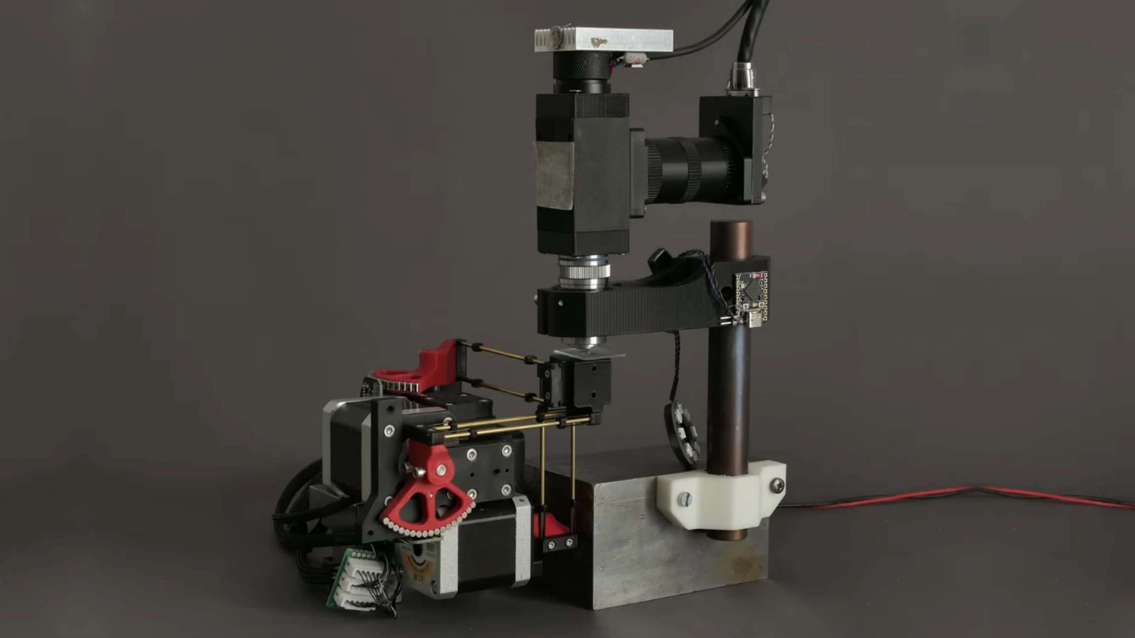 A camera-based microscope is on a stand, looking down towards a slide which is held on a plastic stage. The stage is held in place by three pairs of brass rods, which run to red plastic cranks mounted to three stepper motors. On the opposite side of each crank from the connecting rod is a semicircular array of magnets.