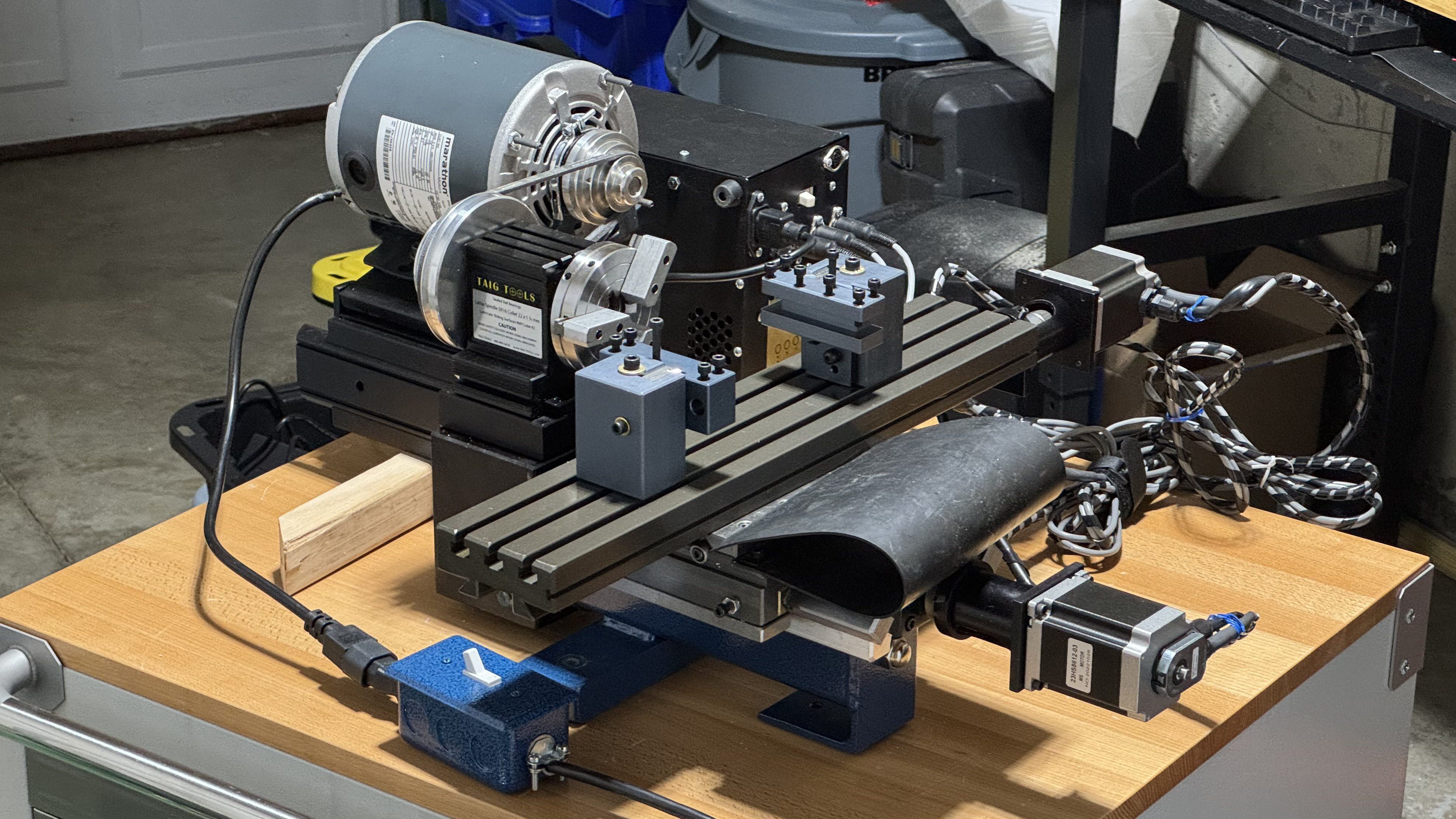 A lathe is shown on a tabletop. Instead of a normal lathe workspace, there is an XY positioning platform in front of the chuck, with two toolposts mounted on the platform. Stepper motors are mounted on the platform to drive it. The lathe has no tailpiece.