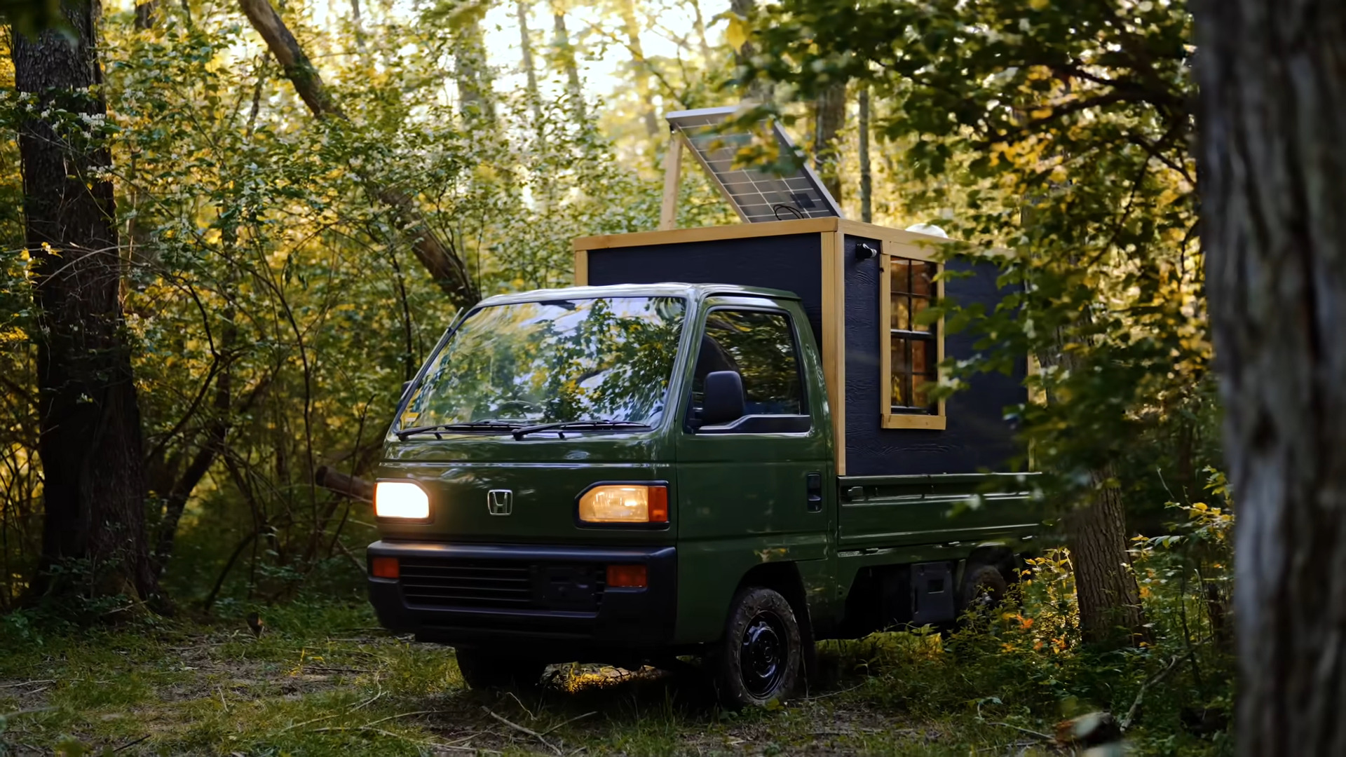 A forest green kei truck sits in an actual forest. In its bed is a dark grey box with a small window, wood trim, and a solar panel tilted on its roof. The headlights are on, hinting at the onset of twilight. You have a sudden urge to go glamping now.