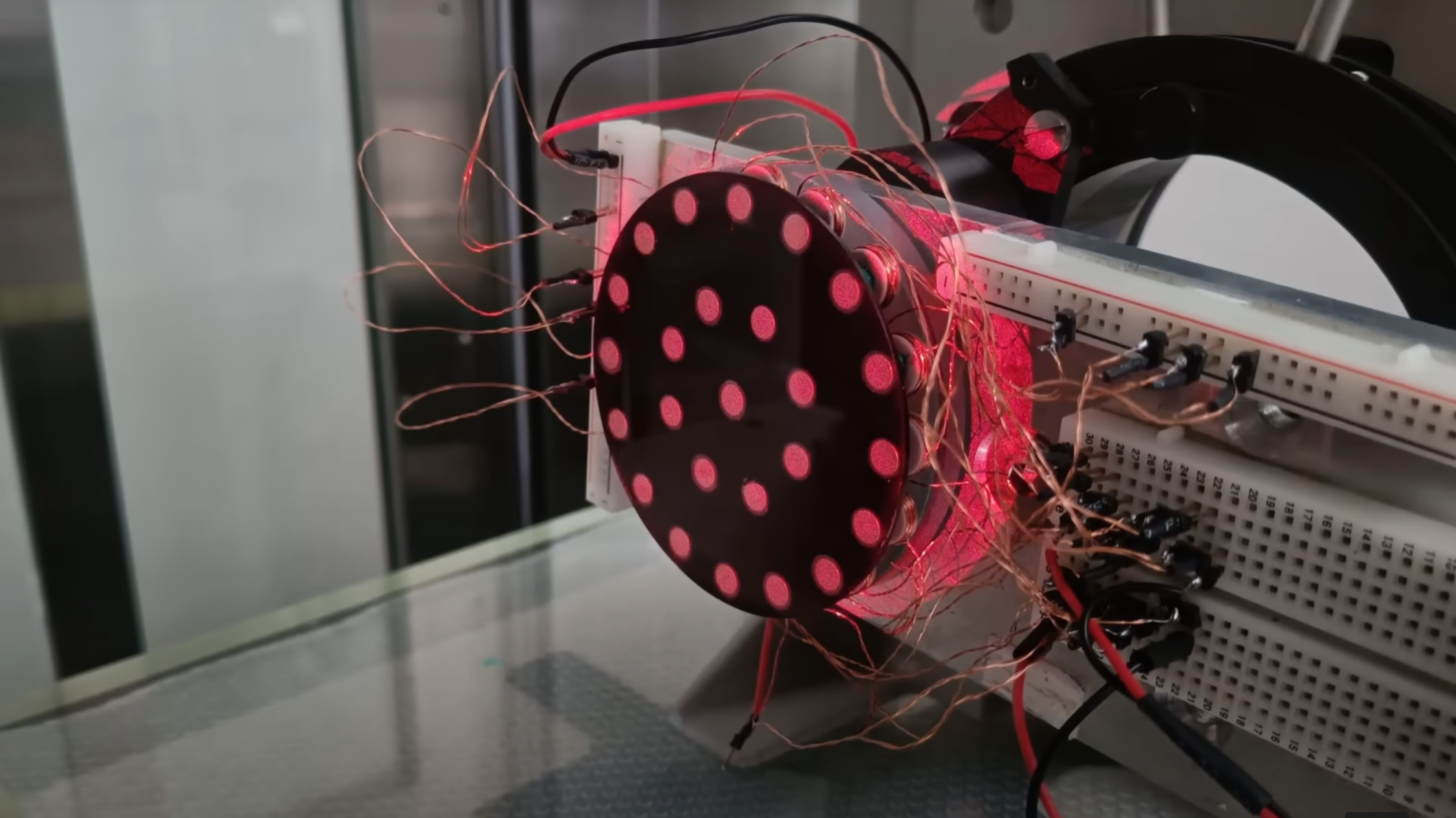A vertically-mounted black disk with a concentric pattern of reflective disks is illuminated under a red light. A large number of copper wires run away from the the disk to a breadboard.