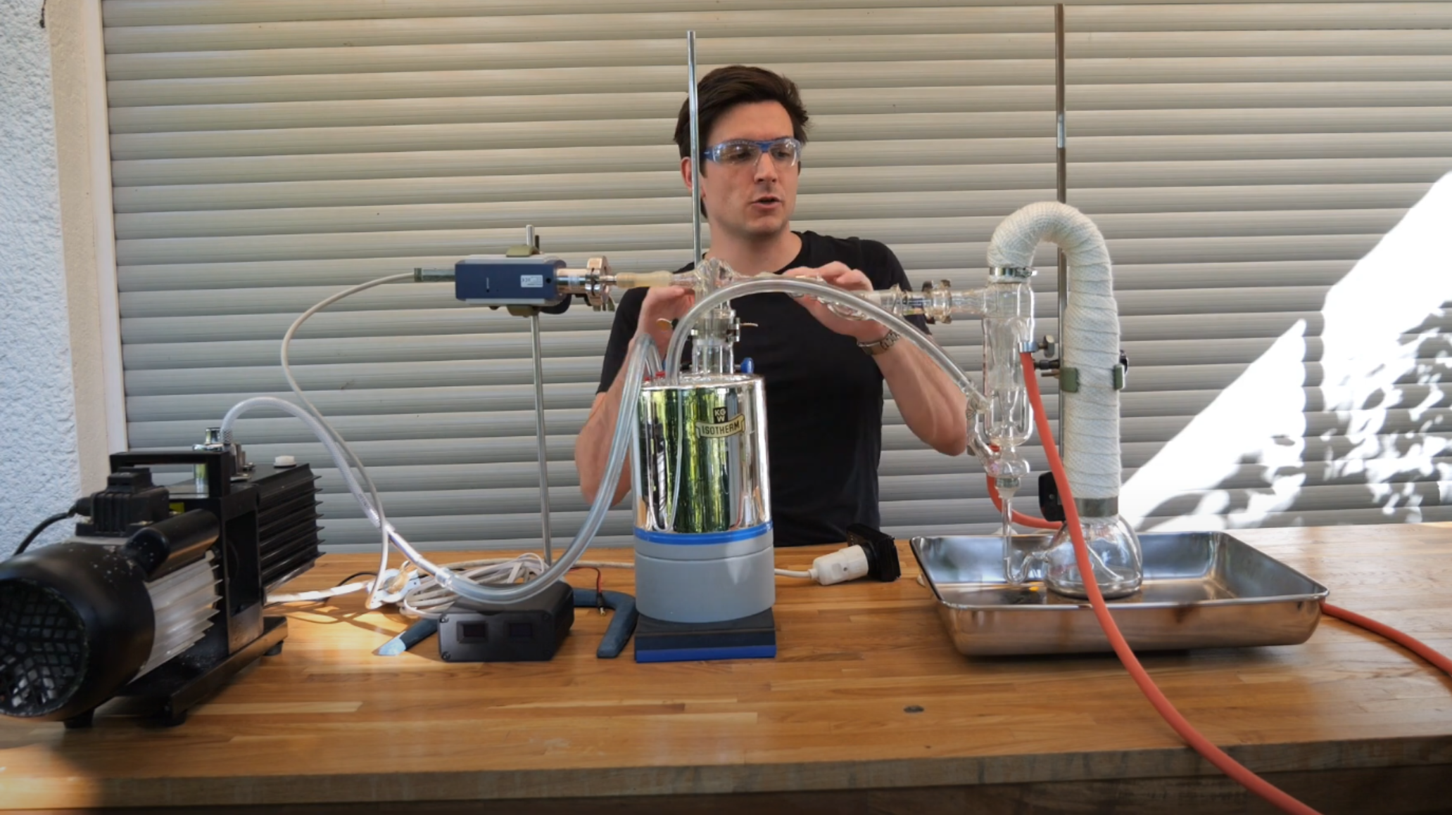 A man is shown behind a table, on which a glass apparatus like a distillation apparatus is set, with outlets leading into a large container in the center of the table, and from there to a pump.