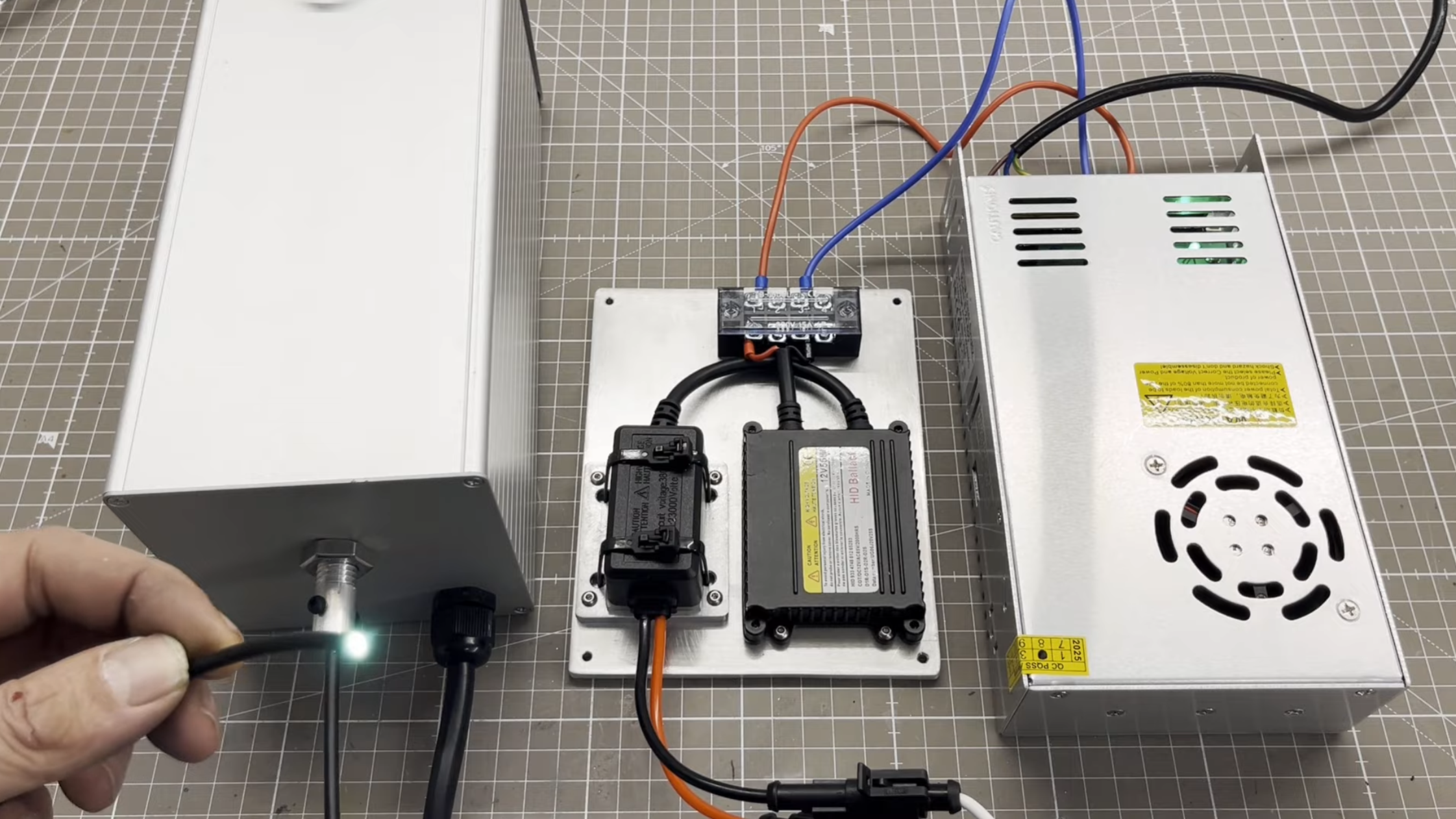 An aluminium box is visible on the left side of the image, with a power supply on the right side, and a lamp ballast in the middle. A man's hand is holding the end of an optical fiber in the lower left corner, and it is emitting a white light.