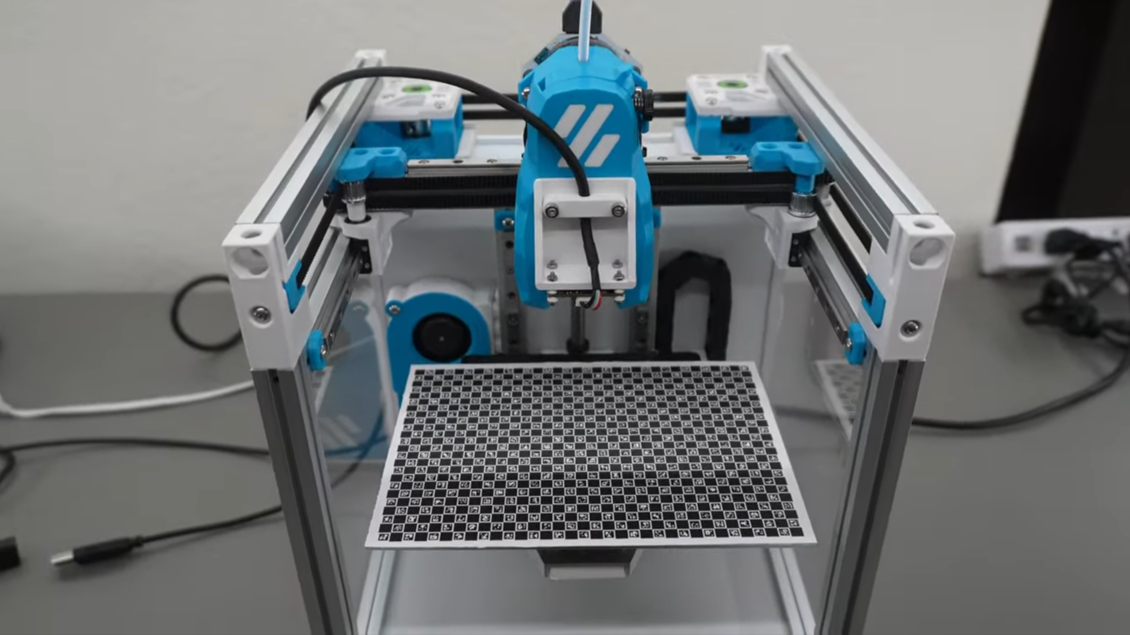 A grey and blue coreXY 3D printer is shown, with a small camera in place of its hotend. On the print bed is a ChArUco pattern, a grid of square tiles containing alternating black fill and printed patterns.