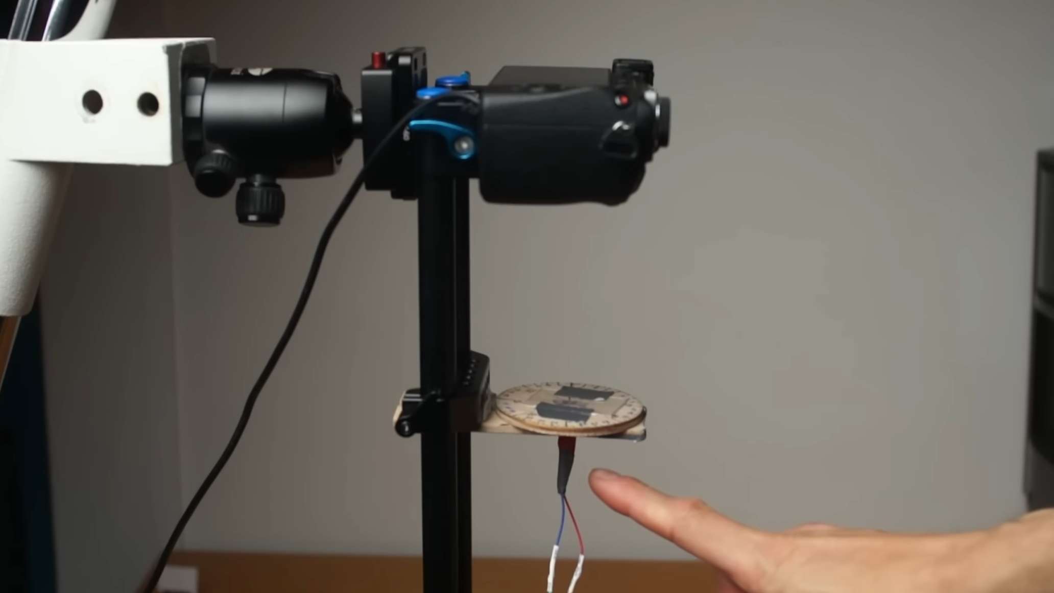 A mirrorless camera is mounted on a stand, facing downwards toward a rotating microscope stage made of wood. A pair of wires come down from the stage, and a man's hand is pointing to the stage.