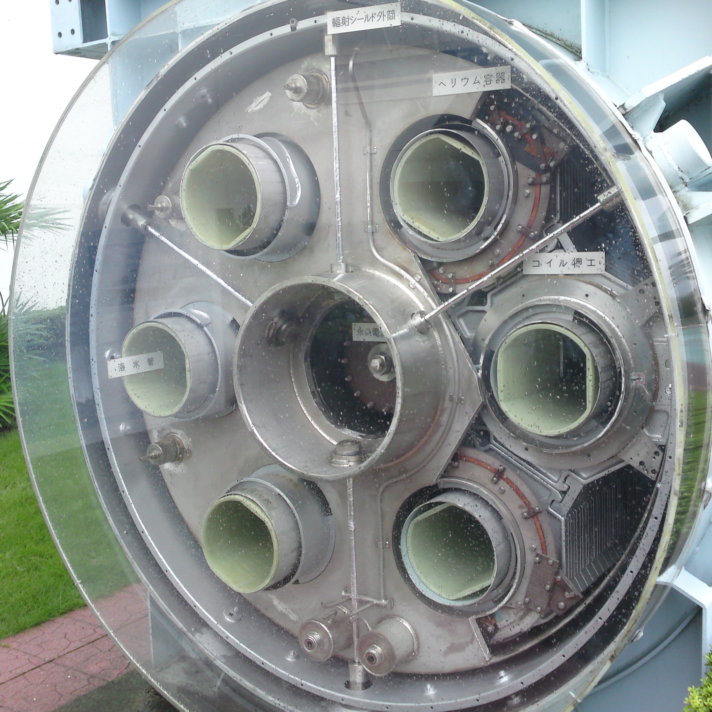 The end of the MHD thruster from the Yamato 1. It has six smaller green tubes surrounding a central circle. All of these pieces are coming through a grey metal fixture that is a circular shape. Small labels are affixed with Japanese writing on them. It is inside a sky blue metal frame.