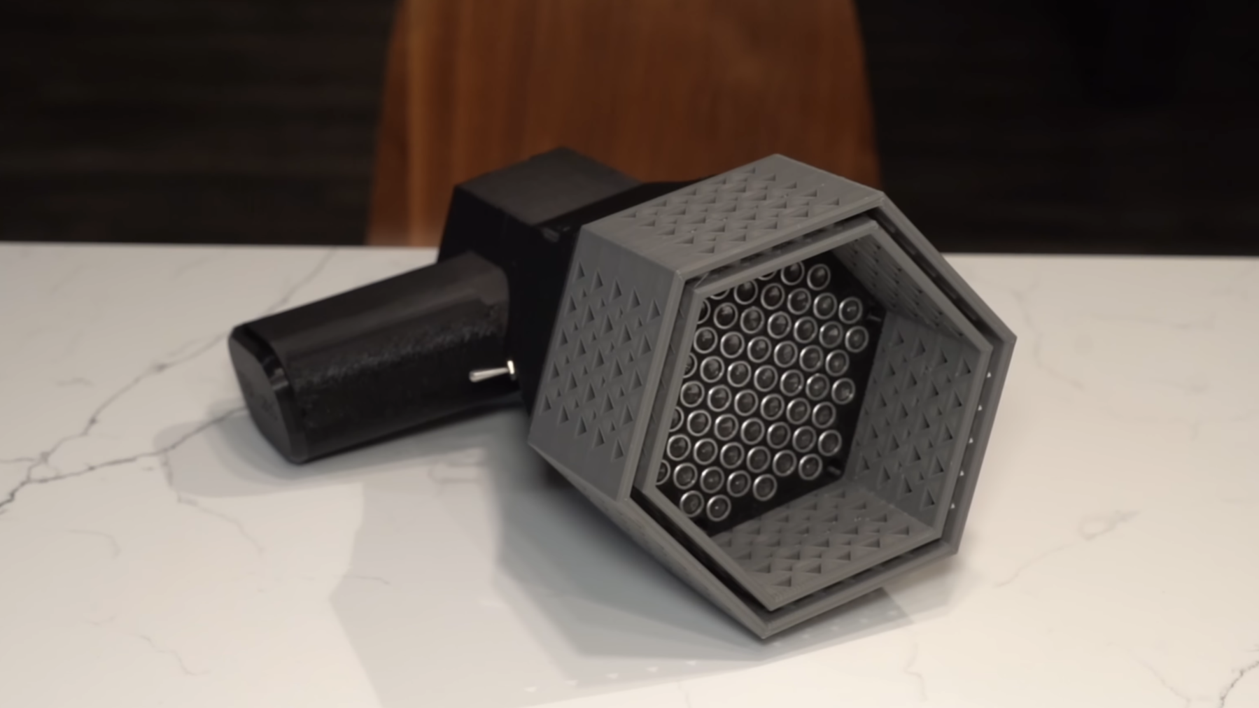 A device rather resembling a megaphone is lying on a table. The handle is made of black plastic. The horn is made of grey plastic, is hexagonal, and is not tapered. At the back of the horn is an array of silver ultrasonic transducers.