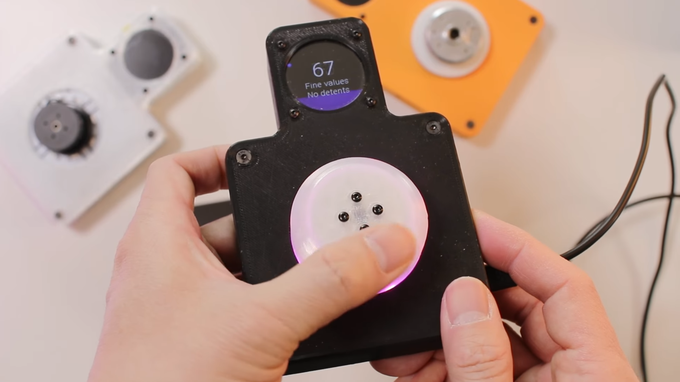 A man's hands are shown holding a black device. A white knob is in the center of the device, and above the knob in a central protrusion from the rest of the device is a small, circular LCD device.