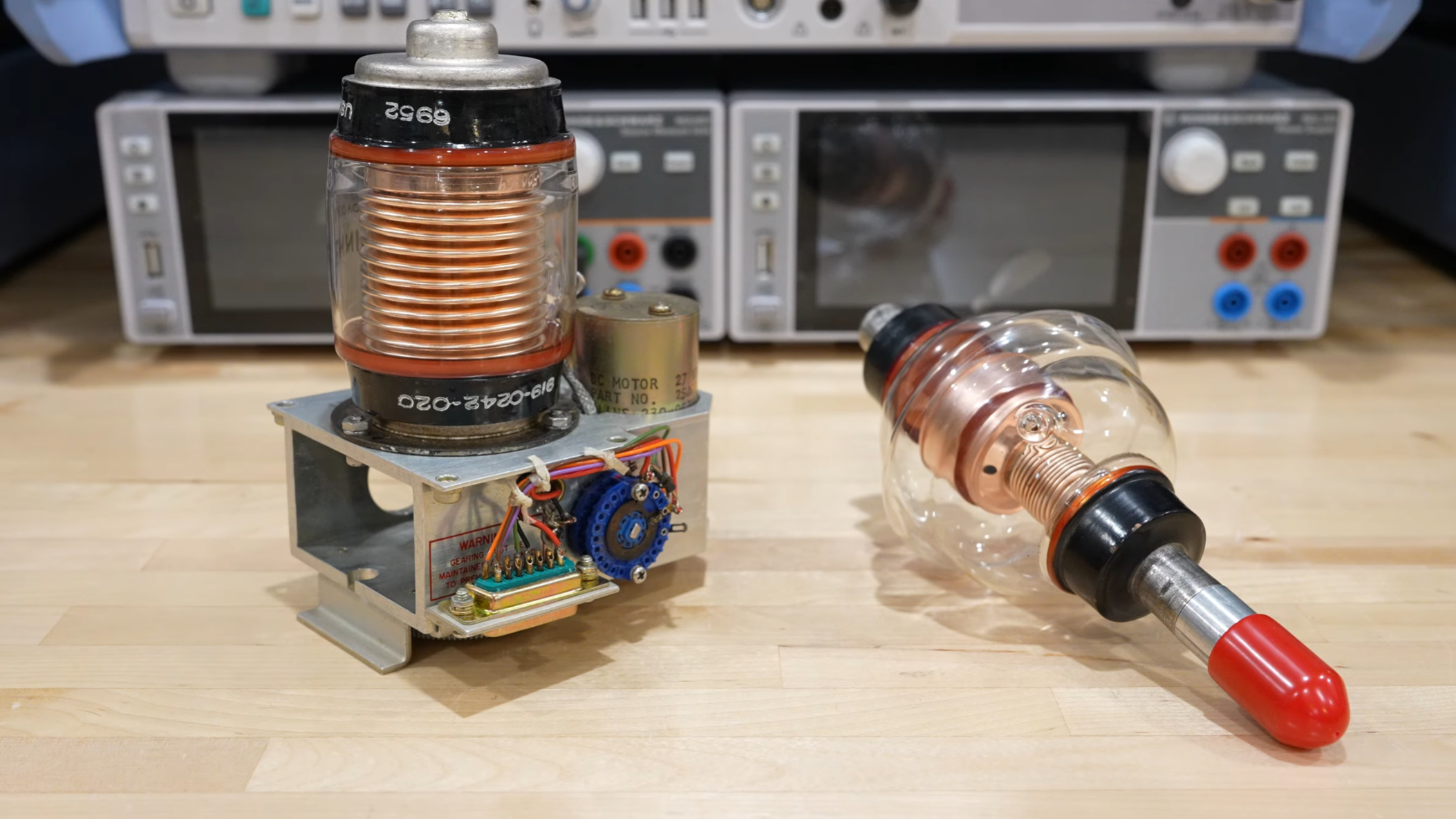 A pair of glass vacuum tubes can be seen on a workbench, each with complex copper structures inside. One is mounted on top of a metal chassis with a motor and some other circuitry visible.