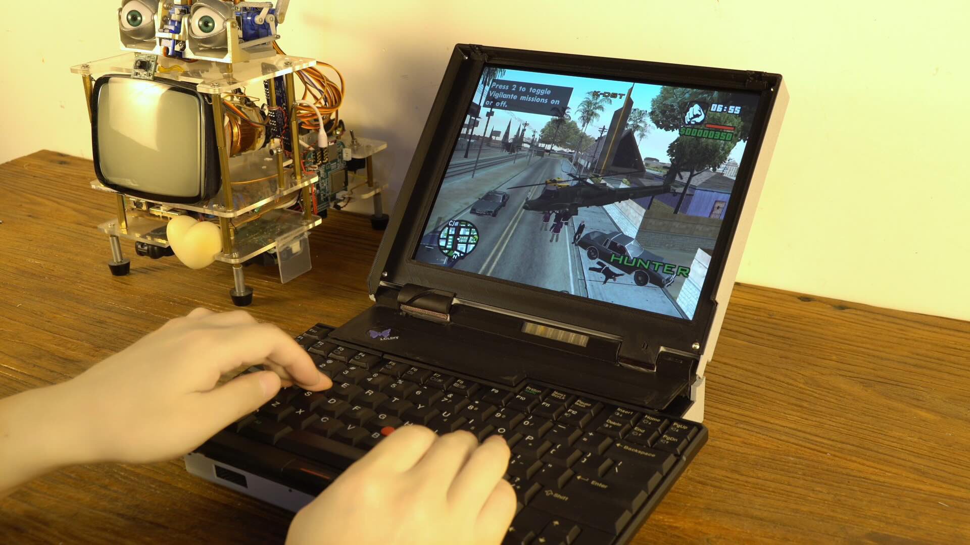 A pair of hands play GTA on a butterfly keyboard-having laptop as a robot looks on.