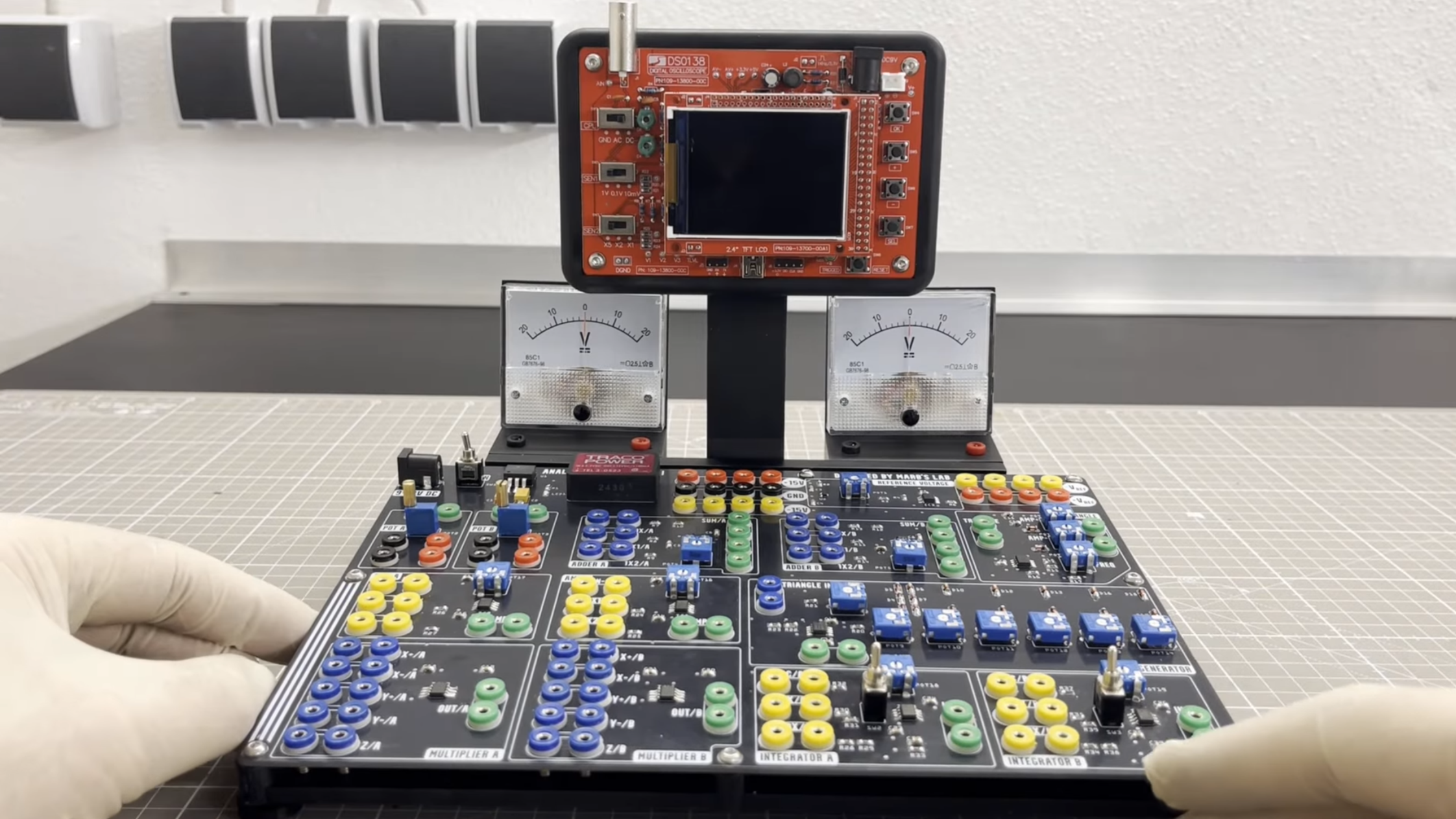 A black circuit board with many colored banana connector plugs in placed on a table. Behind it, two analogue voltage dials stand on either side of an oscilloscope stand. The oscilloscope is a cheap model, with a small LCD display and an exposed red circuit board.