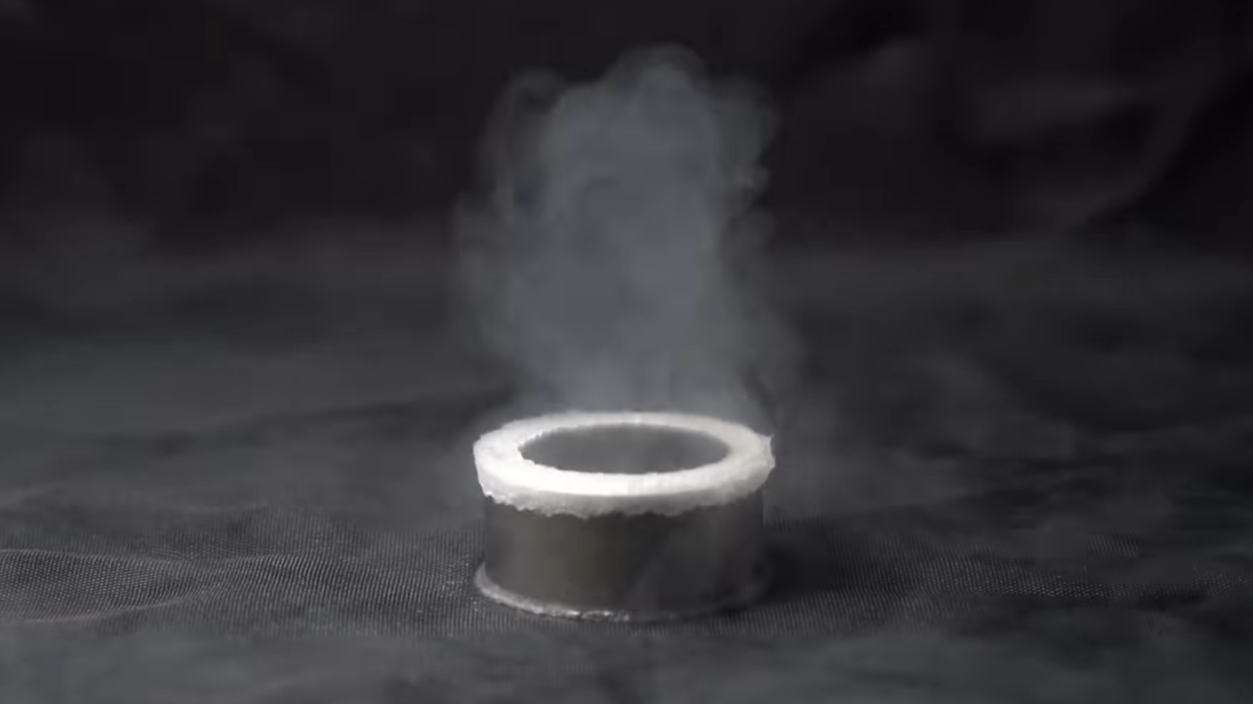 A cylindrical neodymium-iron-boron magnet in shown on a grey background. A white line of boiling liquid surrounds the top edge of the magnet, and a cloud of condensing vapor hangs over it.