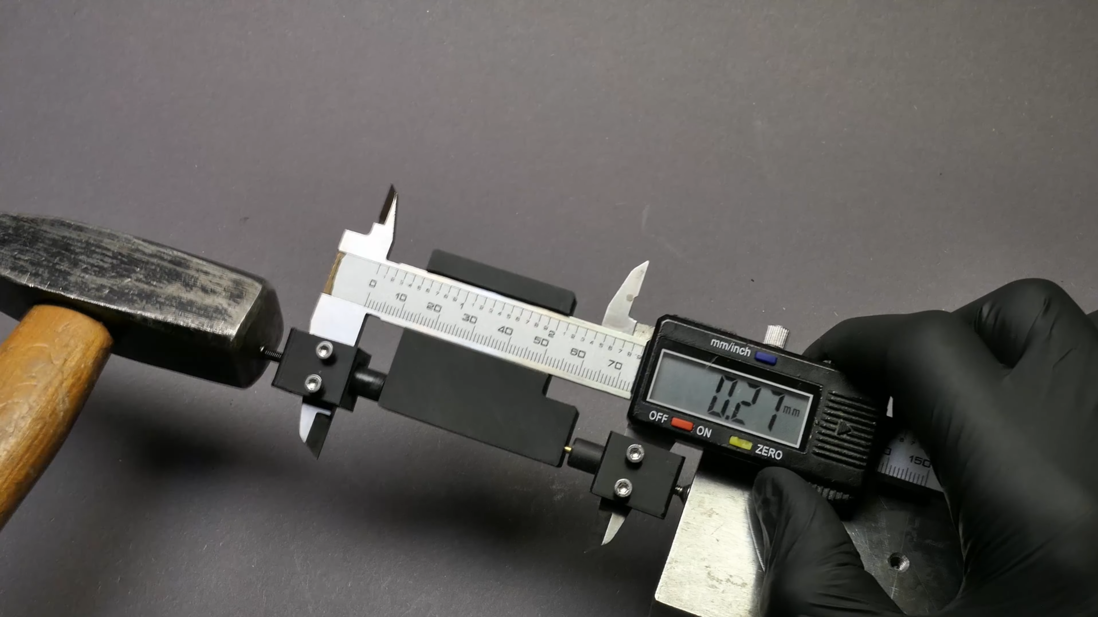 A 3D-printed mechanism is clamped between the jaws of a pair of calipers, which are surrounded by 3D-printed covers. A hammer is resting against one of the jaws, and a man's gloved hand is holding the calipers.