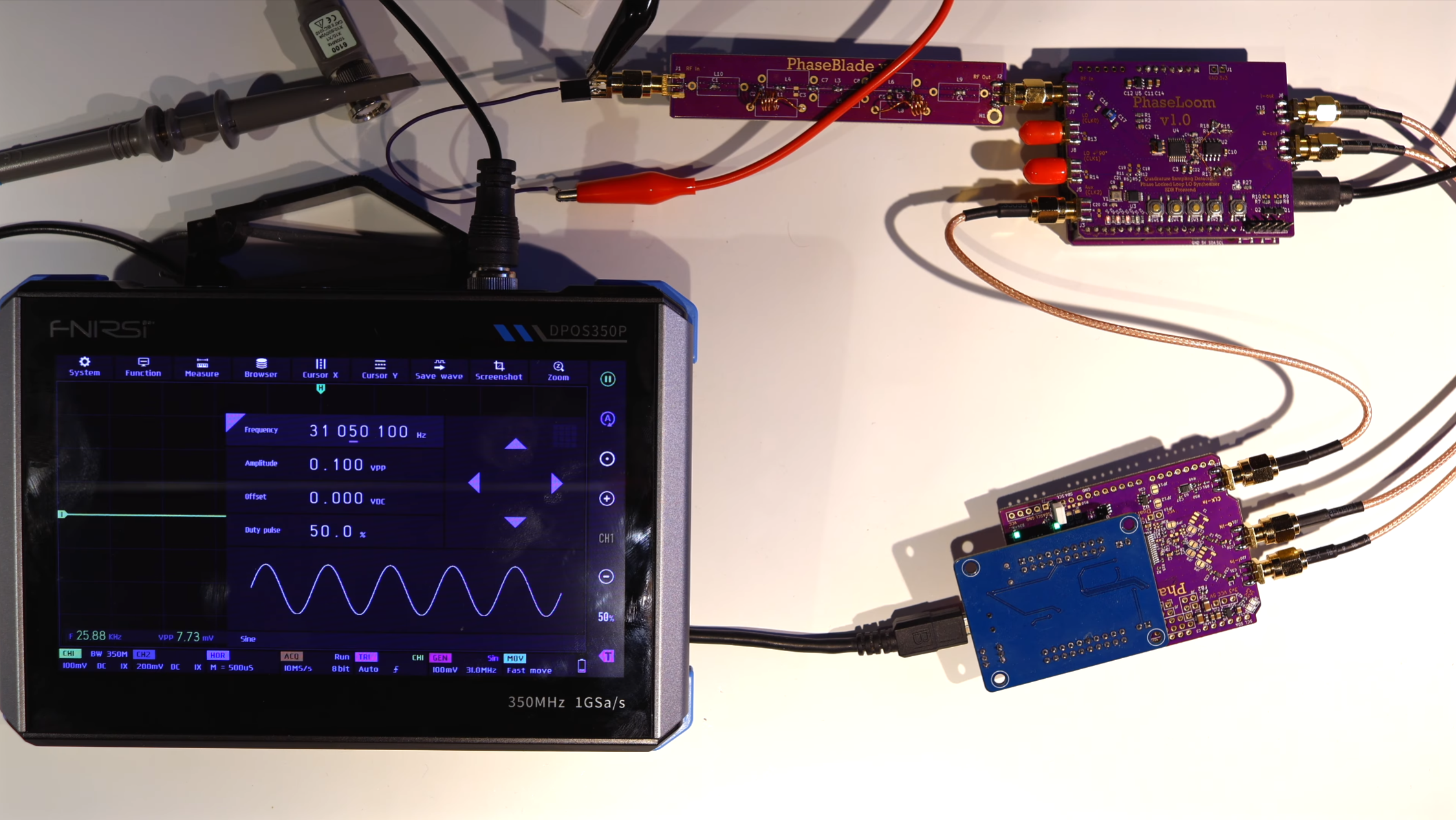 An oscilloscope display is seen in lower left corner. In the rest of the image, two purple circuit boards are connected by SMA RF cables. A wire antenna is connected to one board.