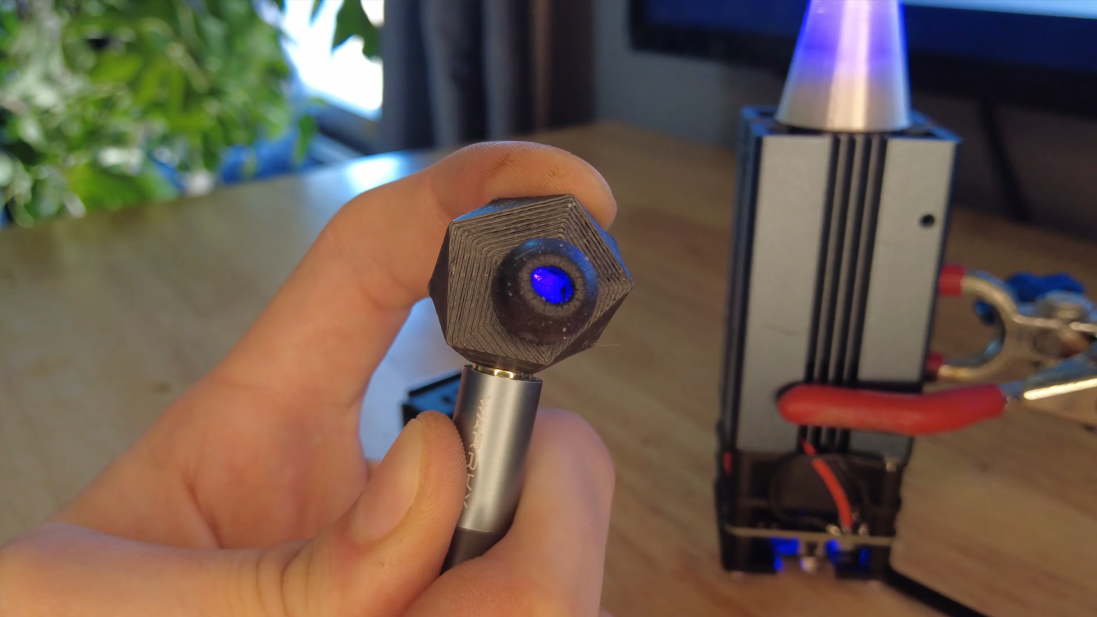 A man's hand is shown holding a 3D-printed structure. The structure is hollow and has a fiber-optic cable leading to it. Blue light shines from a hole in the structure. In the background, a laser module is coupled to a fiber-optic cable.