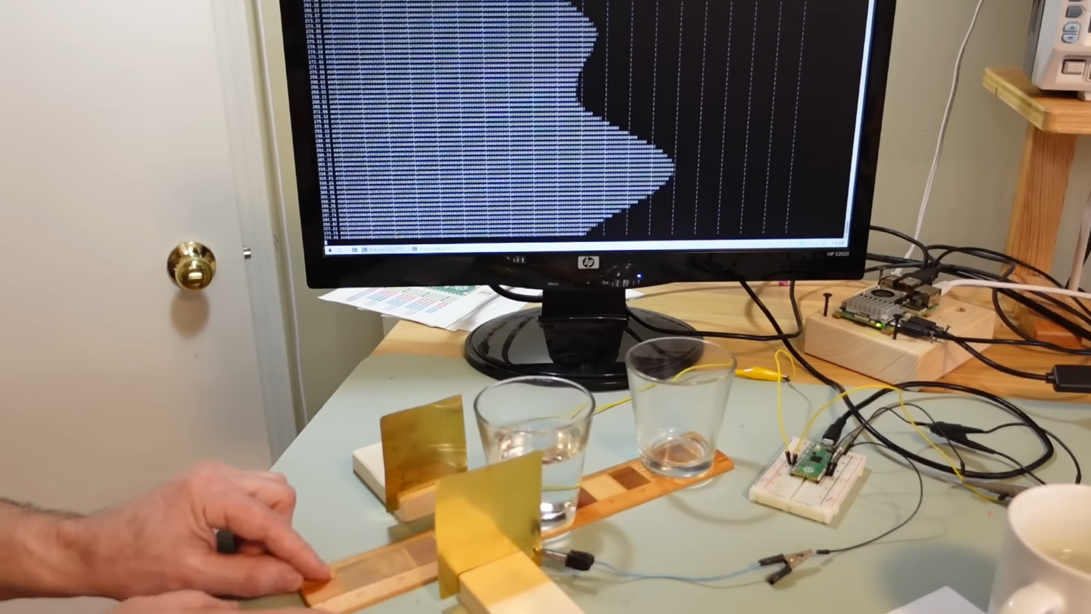 A man's hand is holding a thin wooden board, which runs between two vertical metal plates. On the board are two glasses, one filled with water. A computer screen is behind all this, showing a series of white bars. The series of bars shows two peaks.